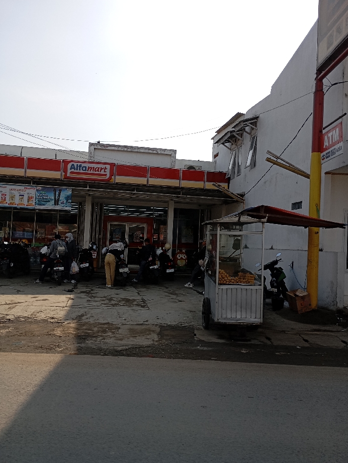 The image captures a daytime street scene in Cipatik, Indonesia, featuring an Alfamart convenience store and an adjacent white two-story building. The Alfamart, characterized by its red and white facade and prominent "Alfamart" sign, has a covered entrance area. Approximately 7-10 individuals are present in front of the store, primarily near a cluster of parked motorcycles, numbering at least five. One person in light-colored pants is observed bending over near a motorcycle. Adjacent to the Alfamart, the white building displays several open windows with awnings and an "ATM" sign on its exterior wall, alongside a red and yellow utility pole. In the foreground, a white mobile vendor cart with a red awning, containing numerous round, yellow-orange items, is positioned on the right. The ground in front of the buildings is a worn, possibly concrete, surface, transitioning to a darker asphalt road in the immediate foreground, which is bisected by a sharp shadow line. The sky is bright and appears overcast or hazy.