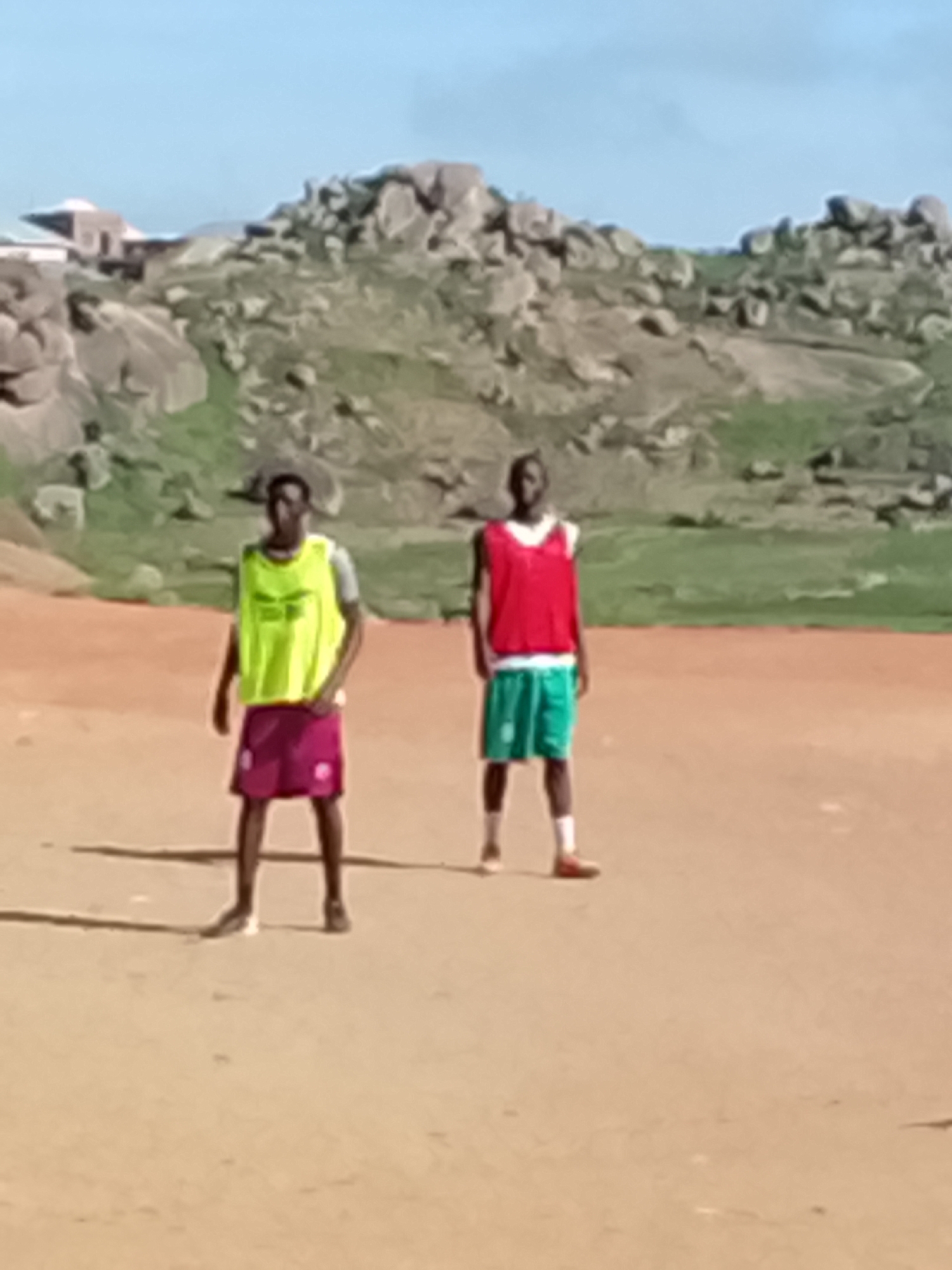 In the city of Jos, Nigeria, on a dirt field, two young men stand facing the camera. The man on the left wears a bright yellow training bib over a grey t-shirt and dark red shorts. He has short dark hair and is looking at the camera. The man on the right wears a red training bib over a white t-shirt and green shorts. He also has short dark hair and is looking forward. Behind them, the landscape is rugged with large rock formations and scattered green vegetation under a clear blue sky. In the background, a building with a conical roof is visible amongst the rocks. The overall impression is of an outdoor training session in a naturally arid environment.
