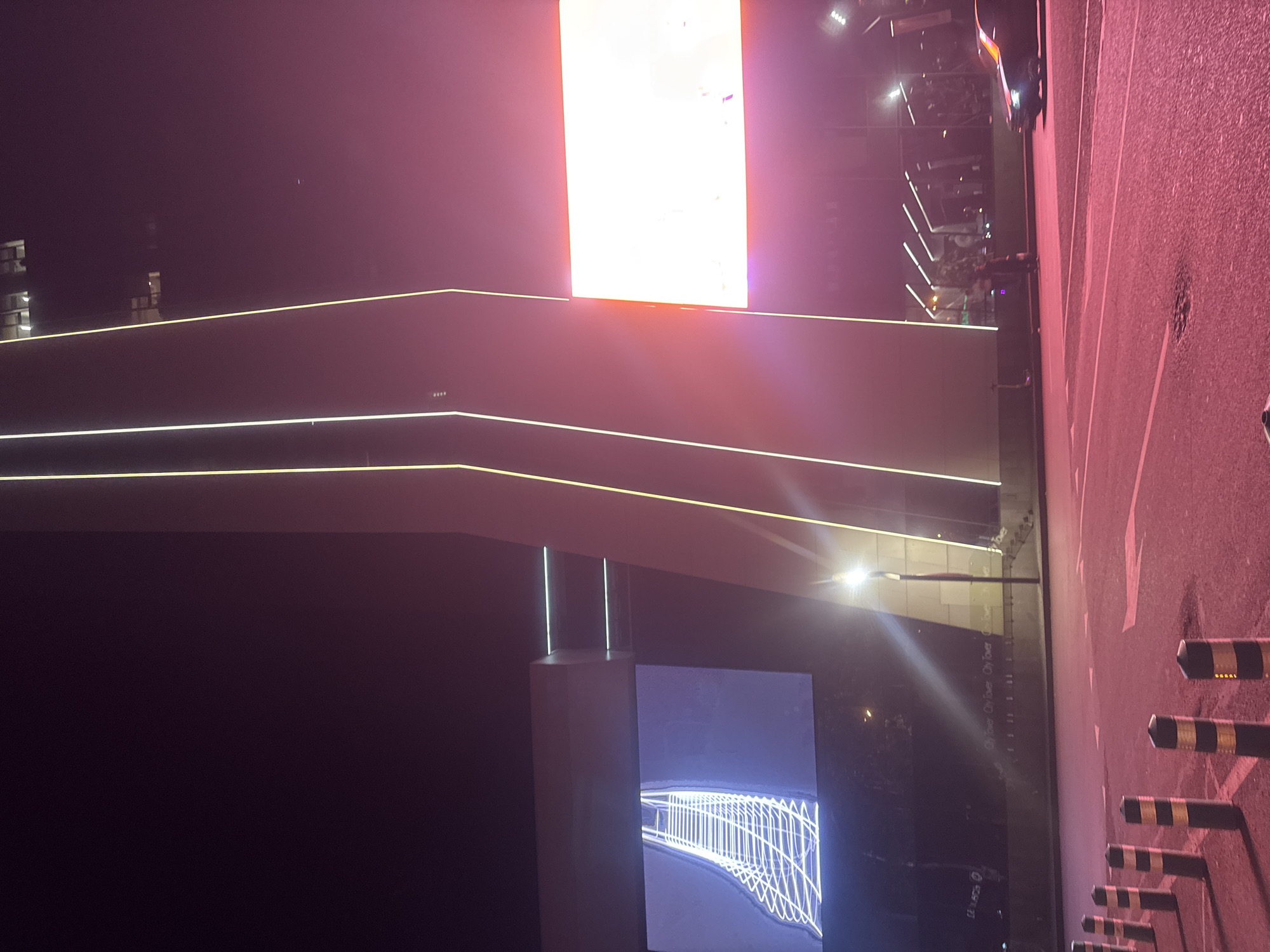 The image depicts a nighttime scene in Tbilisi, Georgia, characterized by artificial lighting and a somewhat hazy or foggy atmosphere, indicated by the soft glow of lights. The composition is dominated by architectural elements and lighting features, suggesting an urban environment.
Prominently featured is a large, bright, rectangular display screen, likely an LED billboard, emitting a strong white light with an orange hue at the bottom, suggesting it's active. Below this, a modern, angular building structure is visible, highlighted by continuous linear lights along its edges and facade. These lights are thin and white, creating a sleek, contemporary aesthetic. Further down, another digital display shows a complex, glowing white lattice pattern against a dark background.
On the right side of the image, a textured asphalt surface, possibly a road or parking lot, is visible, marked with white lines. Several bollards, striped black and yellow, are arranged in a row. A car is partially visible in the background on the right, and a few indistinct figures of people can be seen. The overall mood is one of urban nighttime illumination and modern design, with a touch of mystery due to the low light conditions.
There is some text visible, though it is challenging to read clearly due to the lighting and angle. On the building structure, the words "City Tower" appear repeated. A license plate like string of characters "LL 1573" is also partially visible.