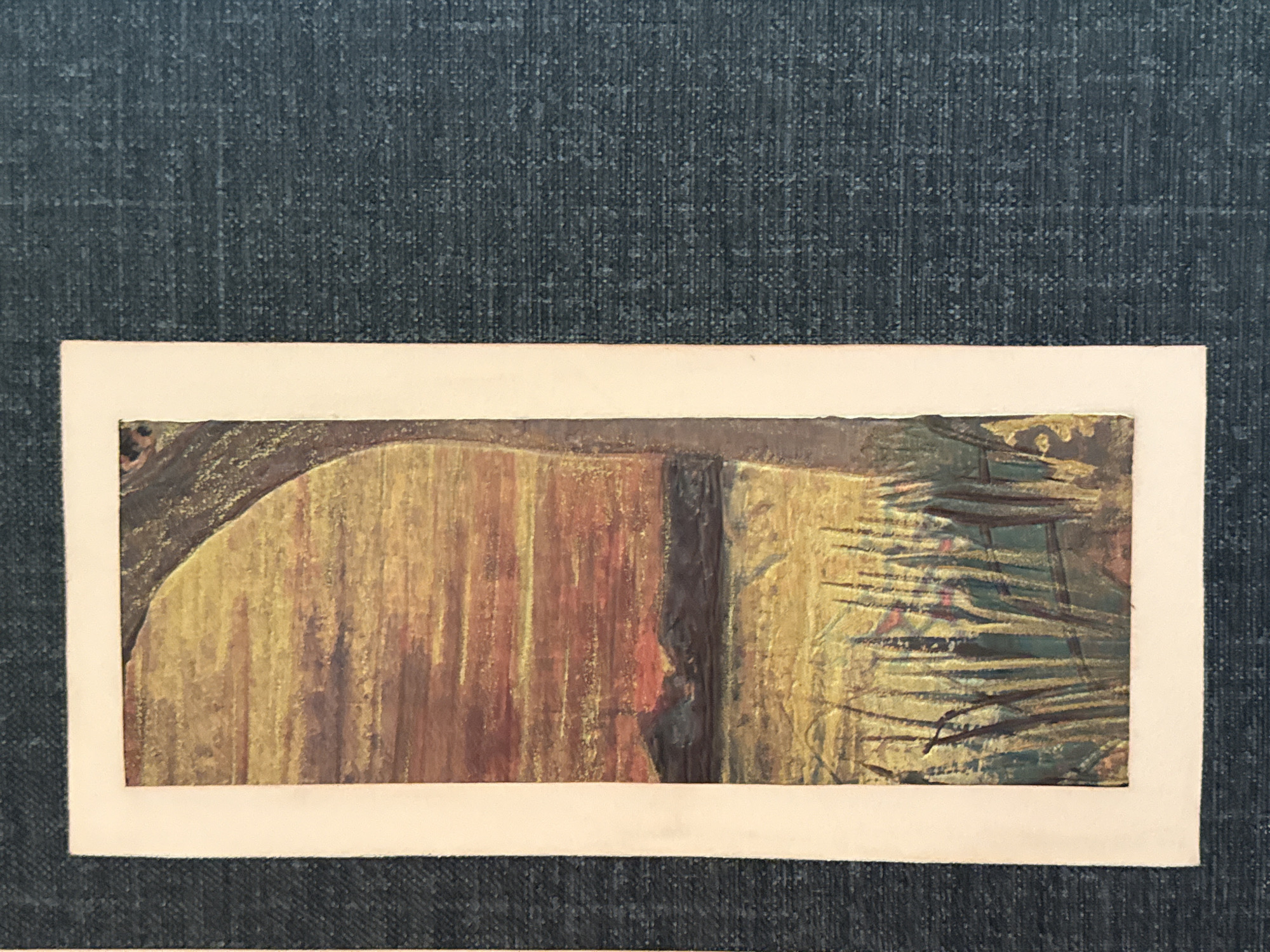 The image is a vertical, close-up shot of a painting depicting a sunset over a body of water with reeds in the foreground. The painting is framed by a white border. The background of the image is a dark, textured fabric, possibly denim. The painting itself features warm tones of orange, yellow, and brown for the sunset and sky, with darker, muted colors for the distant hills and water. The reeds in the foreground are painted in shades of green and brown with hints of red. A large, dark, tree-like shape is visible on the right side of the painting, partially obscuring the sunset. The painting appears to be an oil or acrylic on canvas. There are no people or animals visible in the painting. The location in the city of Galinė, Lithuania is not ascertainable from the image.