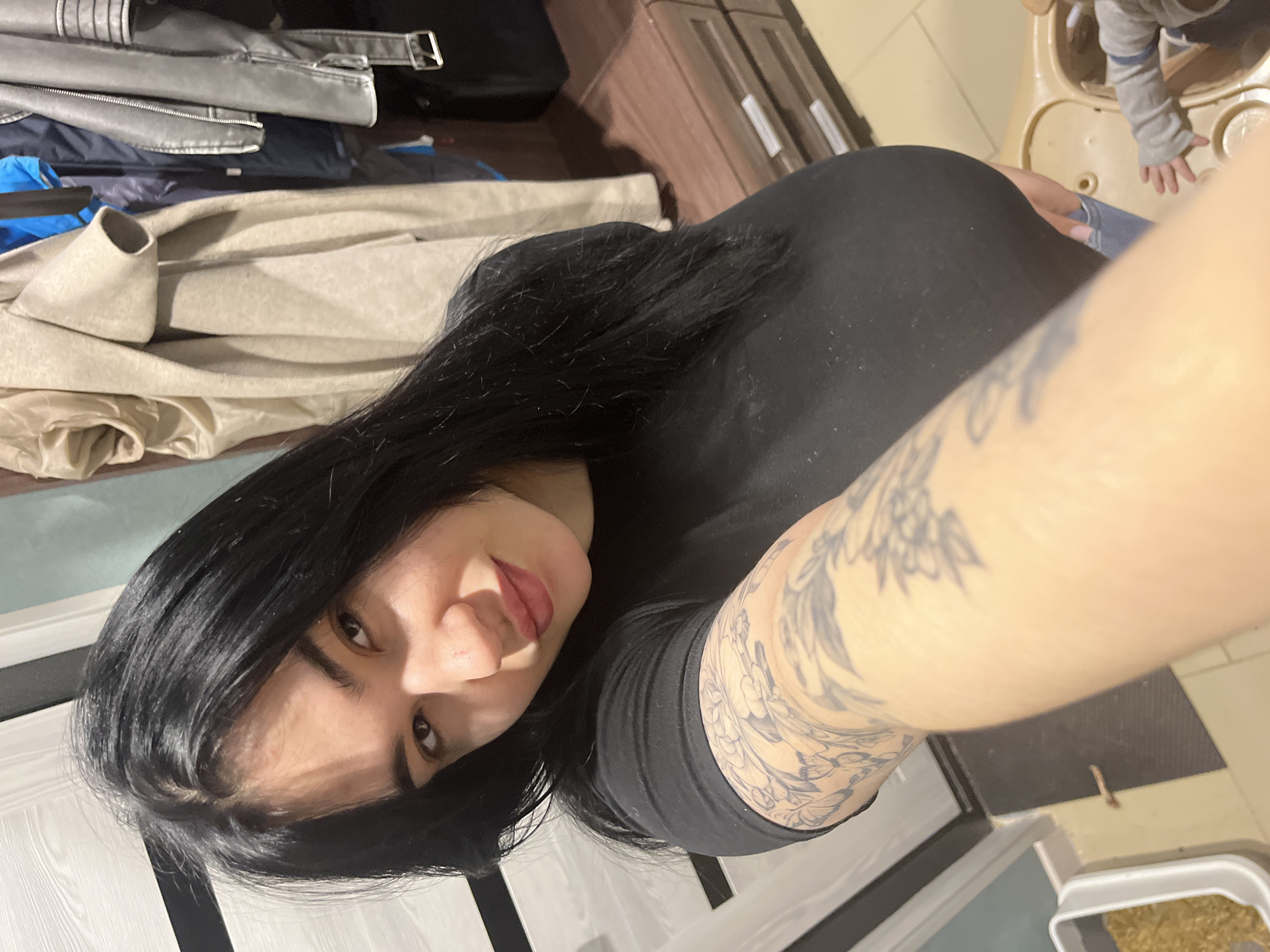 The image displays an adult female taking a selfie within a residential interior in Chaikovsky, Russia. She possesses dark hair, dark eyes, and visible tattoos on her left arm, wearing a black t-shirt and light-colored jeans, looking directly at the camera with a slight smile. The background on the left features a white door with dark accents. To the right, a section of a wardrobe or closet contains hanging garments, including a beige coat and a grey leather-style jacket, positioned above a brown wooden two-drawer dresser with silver handles. In the lower left foreground, a white container filled with granular material, possibly a litter box, rests on a dark mat. The lower right foreground reveals a child's hand, wearing a grey striped sleeve, positioned near a beige plastic child's high chair or activity station.