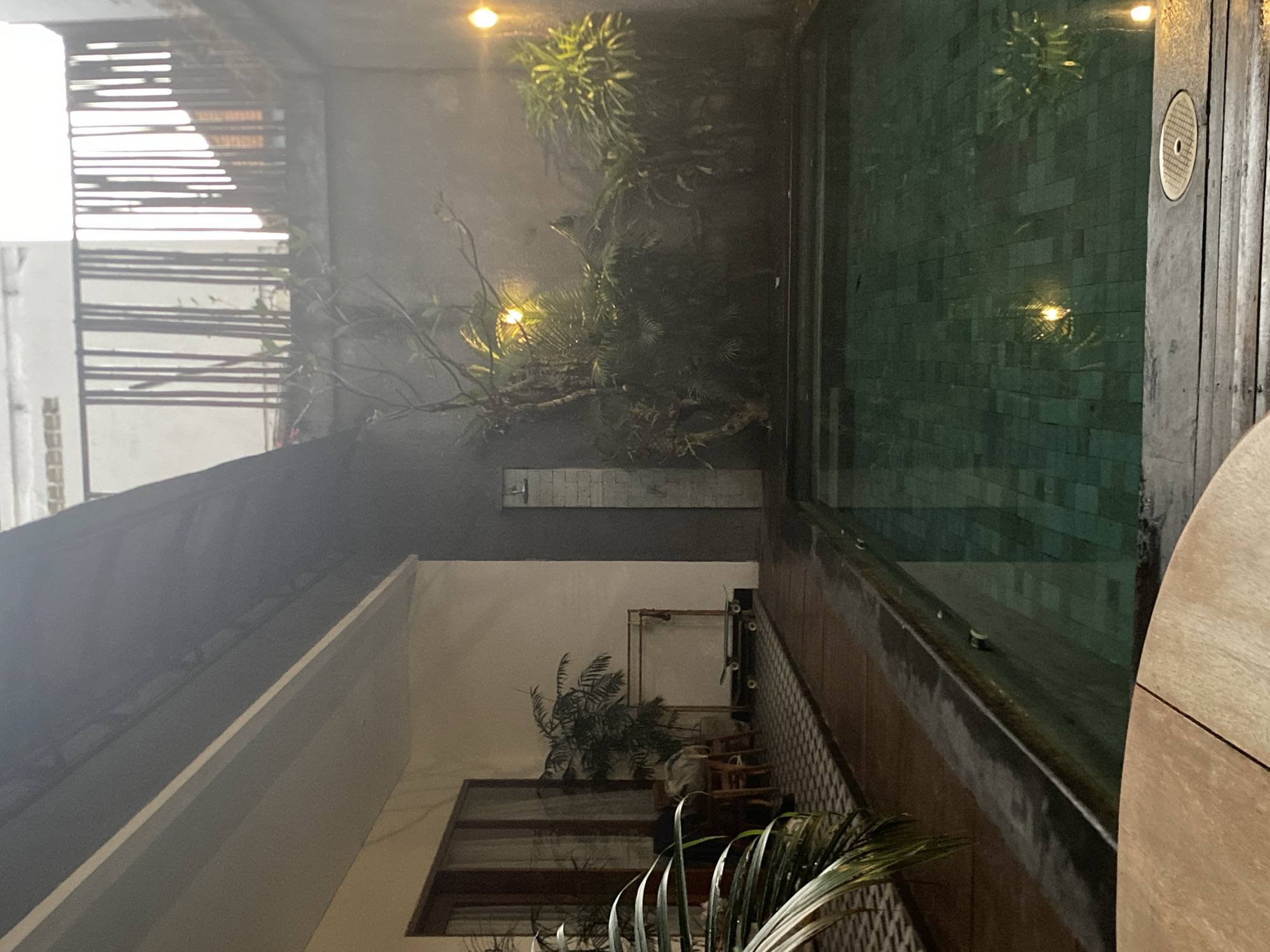 The image depicts a nighttime view of an outdoor swimming pool area in North Kuta, Indonesia. The pool itself is filled with green water, tiled at the bottom, and bordered by wooden decking. Adjacent to the pool is a paved area with a textured pattern, leading to a building with large glass doors and windows. Several potted plants and trees are scattered around the area, some of which are illuminated by yellow outdoor lights, creating a soft glow. A shower head is visible on a brick wall, and a skateboard rests on a clothes rack in the background. The scene is enclosed by a concrete wall and a wooden railing on an upper level, suggesting a private courtyard or resort setting. The lighting and mist create a serene, atmospheric ambiance.