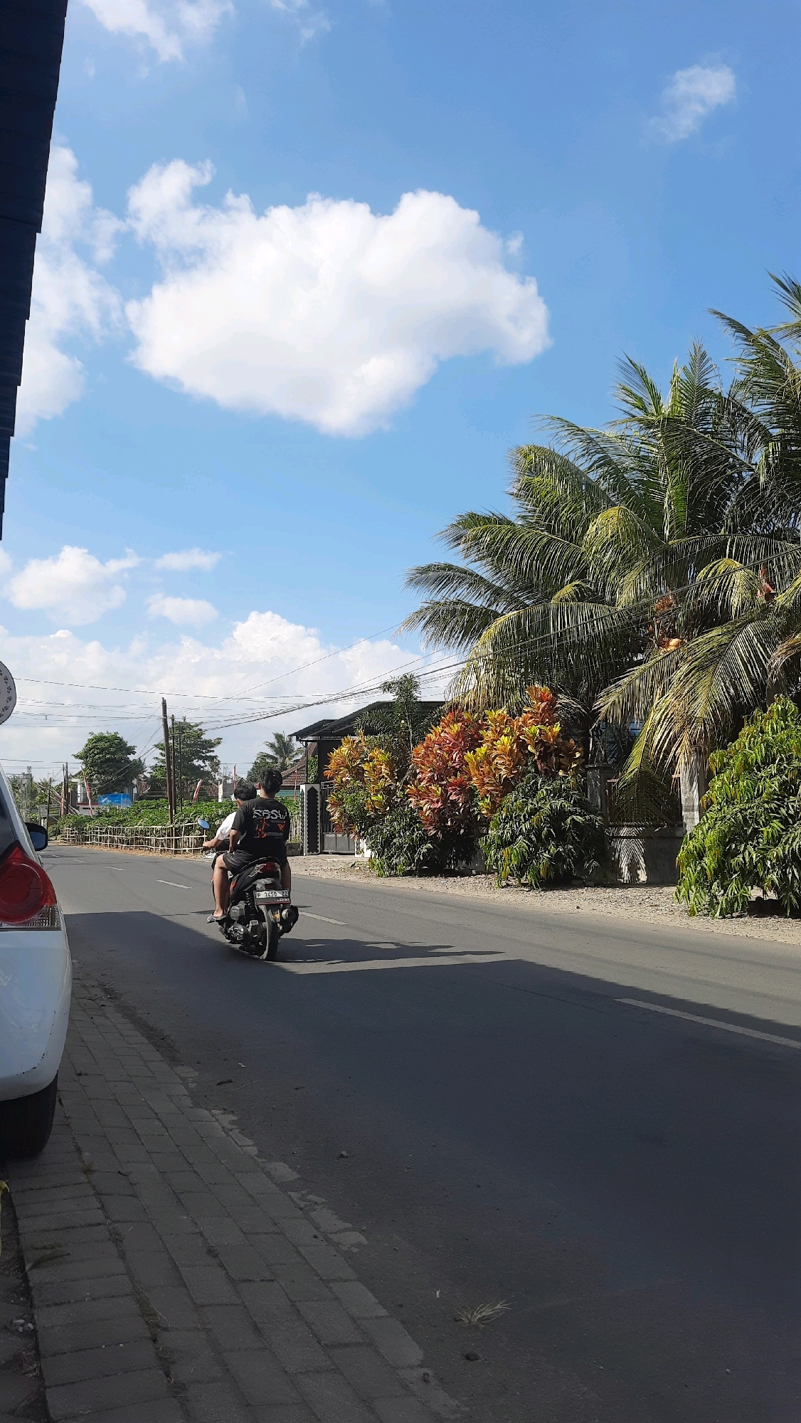This is a full-frame, vertical, eye-level shot taken on a sunny day in Barurejo, Indonesia. The scene depicts a road with a sidewalk on the left and lush tropical vegetation on the right. A white car is parked on the left side of the road, partially visible. In the foreground, a motorcycle carrying two people is traveling away from the camera on the right side of the road. The person in the back is wearing a black t-shirt with white lettering. The road is paved, and the sidewalk is made of interlocking bricks. Power lines are visible overhead, stretching across the frame. Behind the road, there are buildings and more trees, including palm trees. The sky is a clear blue with scattered white clouds. The overall atmosphere is one of a typical day in a tropical urban or suburban environment.