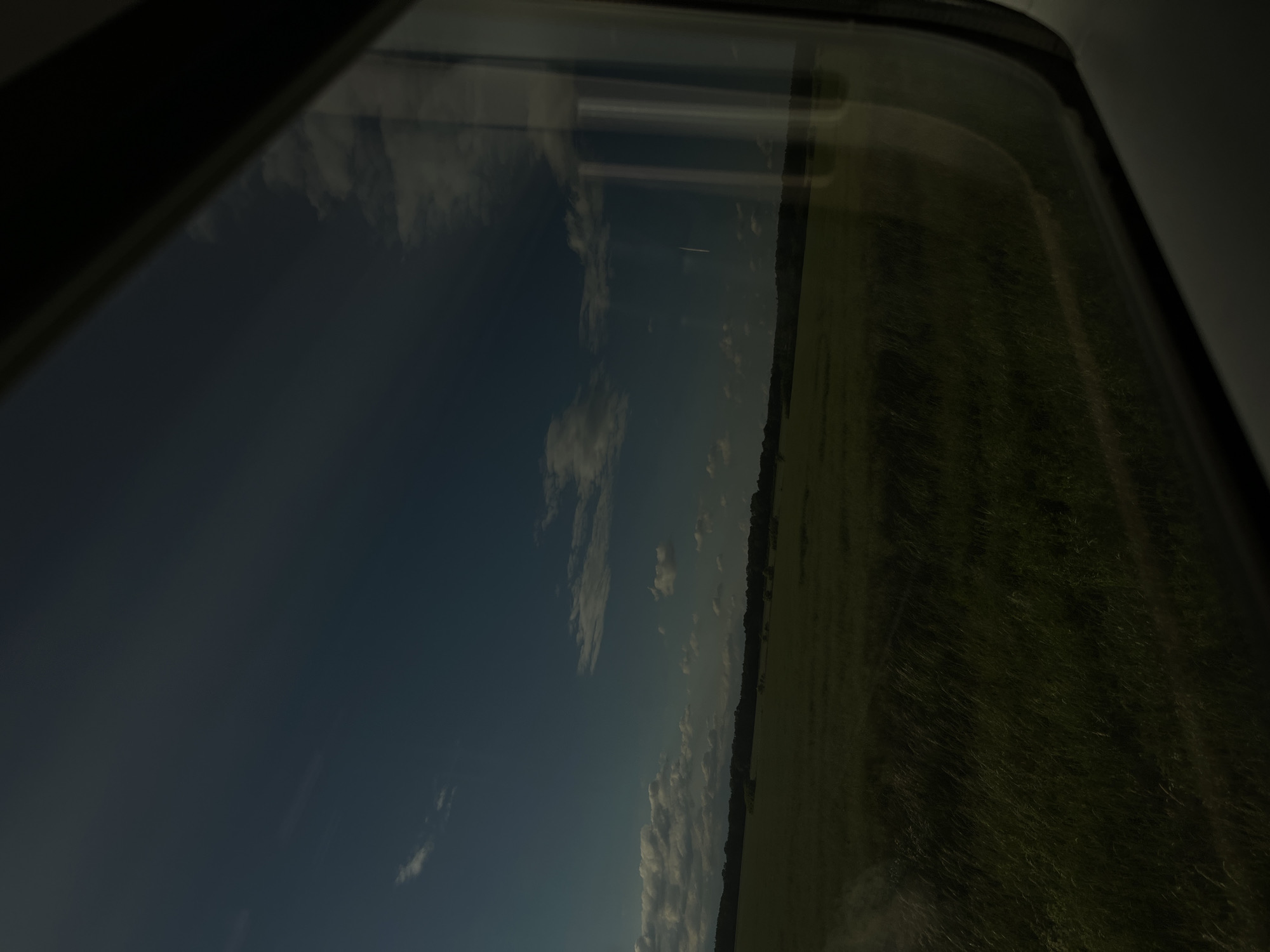 The image shows a view from a train window overlooking a green field and a blue sky with some clouds. The reflection on the window shows some vertical lines, possibly from the train's interior. The field appears to be covered in grass and extends towards a line of trees in the distance. The sky is a clear blue with scattered white clouds, some of which are fluffy and others are wispy. The scene suggests a rural or semi-rural landscape. There are no people or specific landmarks visible that would allow for pinpointing a location within Wittenberg, Germany.