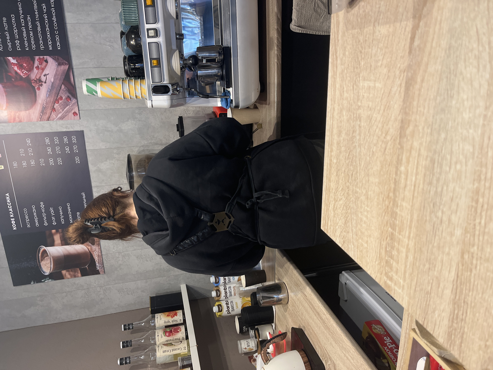 A barista, seen from the rear, is working at an espresso machine in a cafe, as indicated by the menu on the wall listing various coffee drinks. Syrup bottles and plant-based milk containers are visible on the counter. The cafe may be in Chaikovsky, Russia, inferred from the menu text in Russian. A box of Orion Choco Pie is visible in a shelf under the counter.