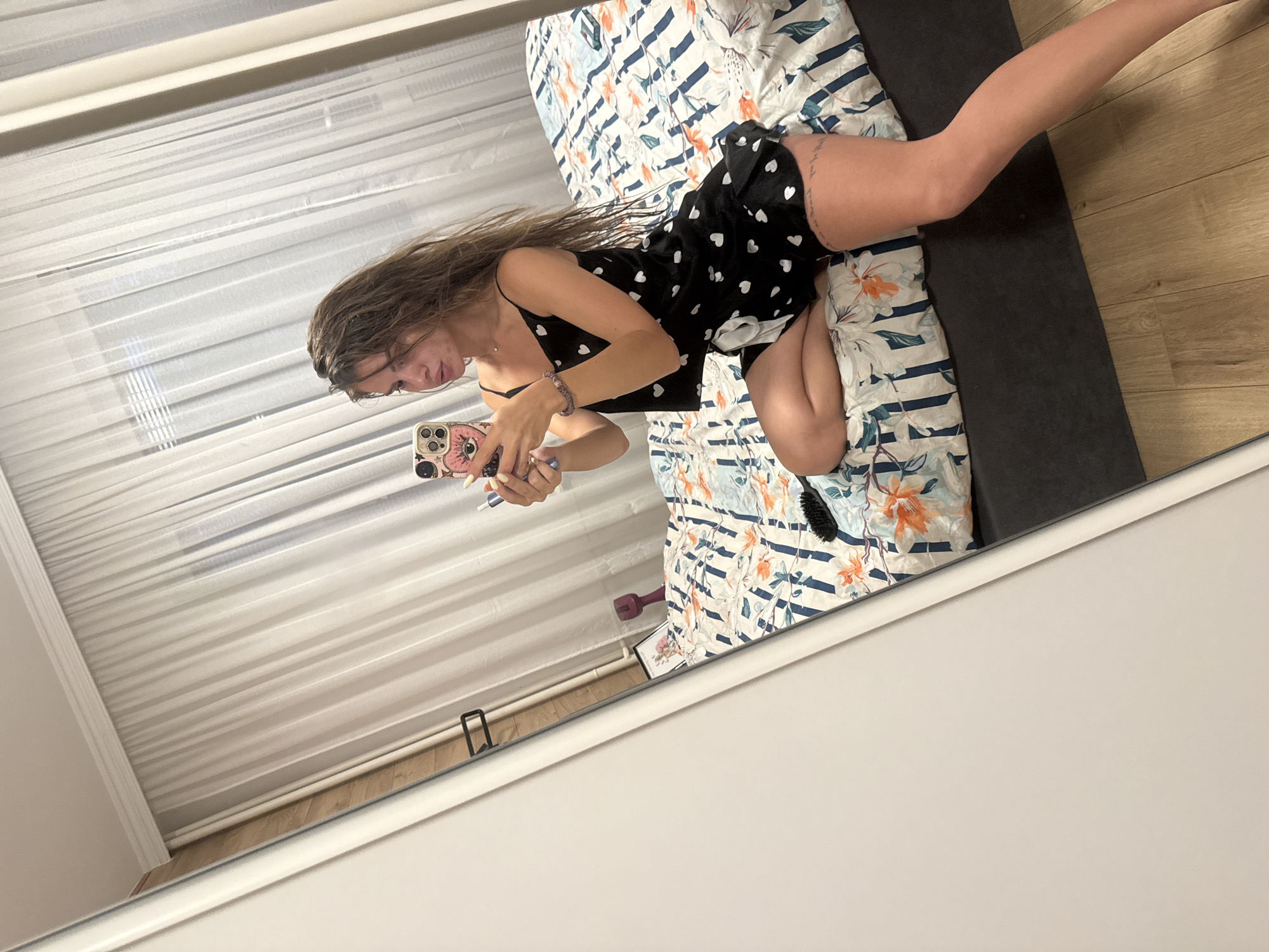 A young woman with long, wet, brown hair is taking a mirror selfie in a bedroom. She is wearing a black pajama set with white hearts and sitting on a bed with floral patterned bedding. She is holding a smartphone with a pink case and a blue lipstick. Her left leg is bent, and her right leg is extended. A black hairbrush is on the bed next to her. The room has white sheer curtains and a wooden floor. There is a white wardrobe to the left of the frame. The image was taken inside an apartment in Plovdiv, Bulgaria.