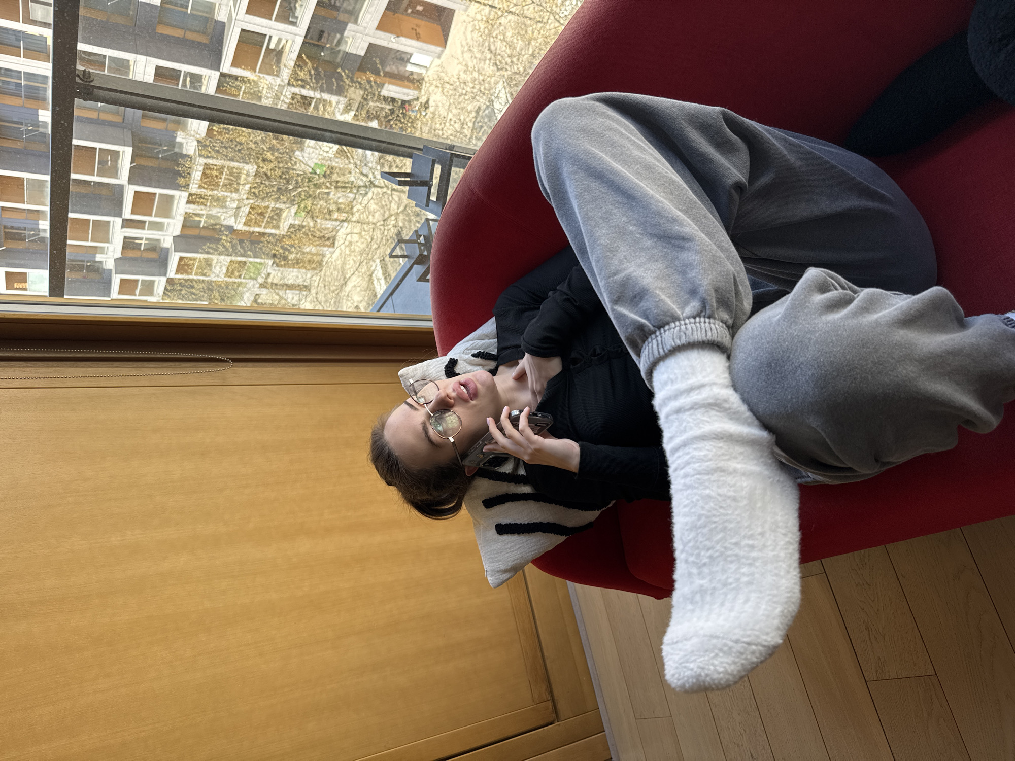 A young person reclines on a red armchair indoors, next to a large window overlooking an urban landscape in Warsaw, Poland. They are wearing a black long-sleeved top and grey sweatpants, with a white fluffy sock on their visible foot. Their head rests on a white and black striped cushion. The person wears round, metallic-framed glasses and holds a smartphone to their ear or near their face, appearing engaged in a call or listening intently, with their mouth slightly open.
The indoor environment features a light wooden floor and a wooden panel or blind beneath the window. Outside, multiple multi-story apartment buildings with visible windows and balconies are seen under natural daylight, suggesting a clear day in late winter or early spring. Bare trees with delicate branches are also visible, hinting at the season. The overall scene depicts a relaxed, casual moment in a residential setting. No visible text is present.