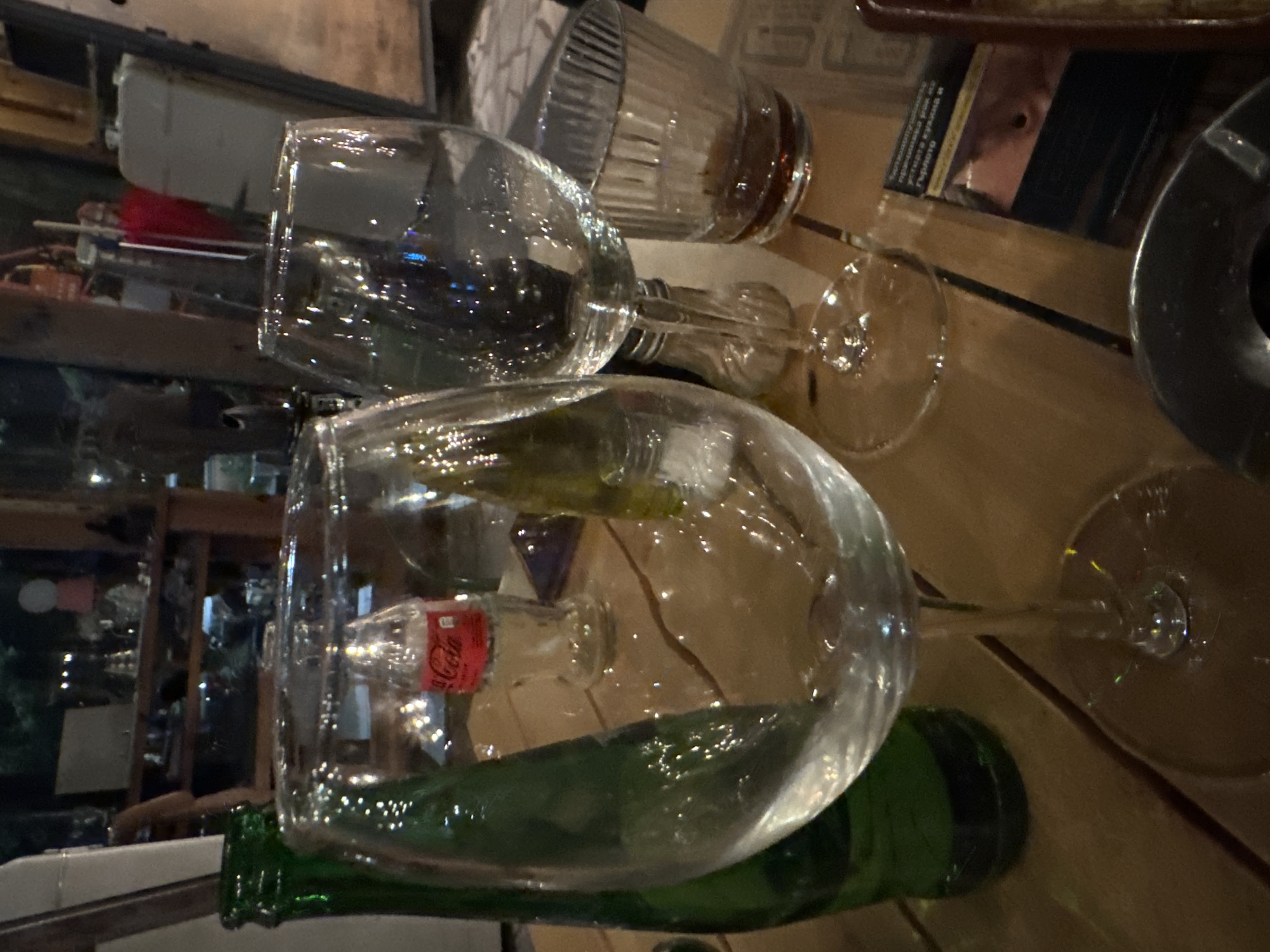 The image shows a table at an indoor location, possibly a restaurant or bar. Several objects are visible on the table, including a green bottle, a Coca-Cola bottle, two wine glasses, a ribbed glass, and an ashtray. The reflective nature of the glasses indicates they are clean and well-maintained. The environment is dimly lit with other furniture and objects visible in the background, suggesting a public space. While specific landmarks are not discernible, the location is indicated as Sofia, Bulgaria.
