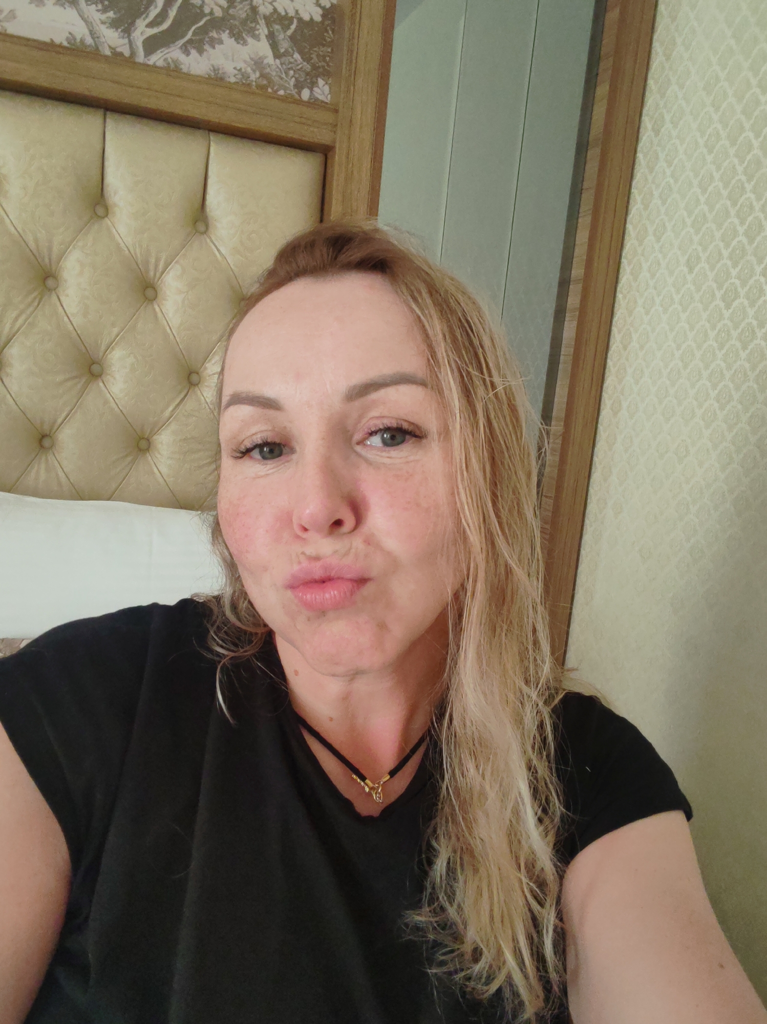 A medium-shot selfie depicts a fair-skinned woman with light brown hair. She is positioned in front of a headboard, with wallpaper visible on the right. She is wearing a black t-shirt and a delicate necklace with a pendant. She has a facial expression with pursed lips. The background suggests an interior setting. It is not possible to verify or determine any city information.