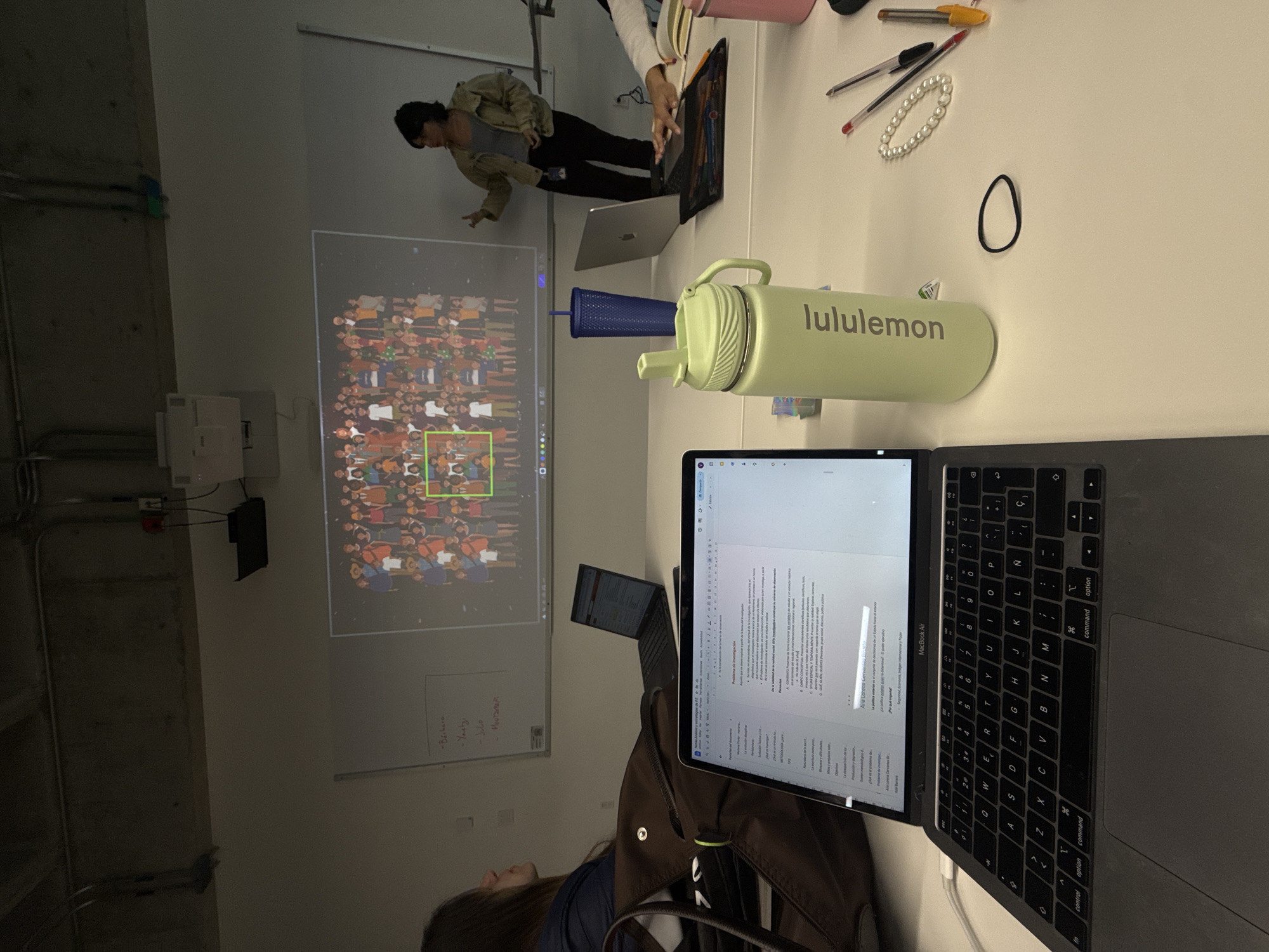 A photograph shows an indoor scene, likely a meeting or classroom environment. A projector is casting an image of a crowd onto a wall, and some people are looking at the screen. One person appears to be presenting in front of the projected image. There's a table on the right side of the frame with various objects: laptops, pens, a bracelet, a hair tie, and a Lululemon water bottle. One laptop is open, displaying text on the screen. A backpack is lying next to the laptop. A person is seated at the table. The image does not provide enough information to determine the location in Mexico City.