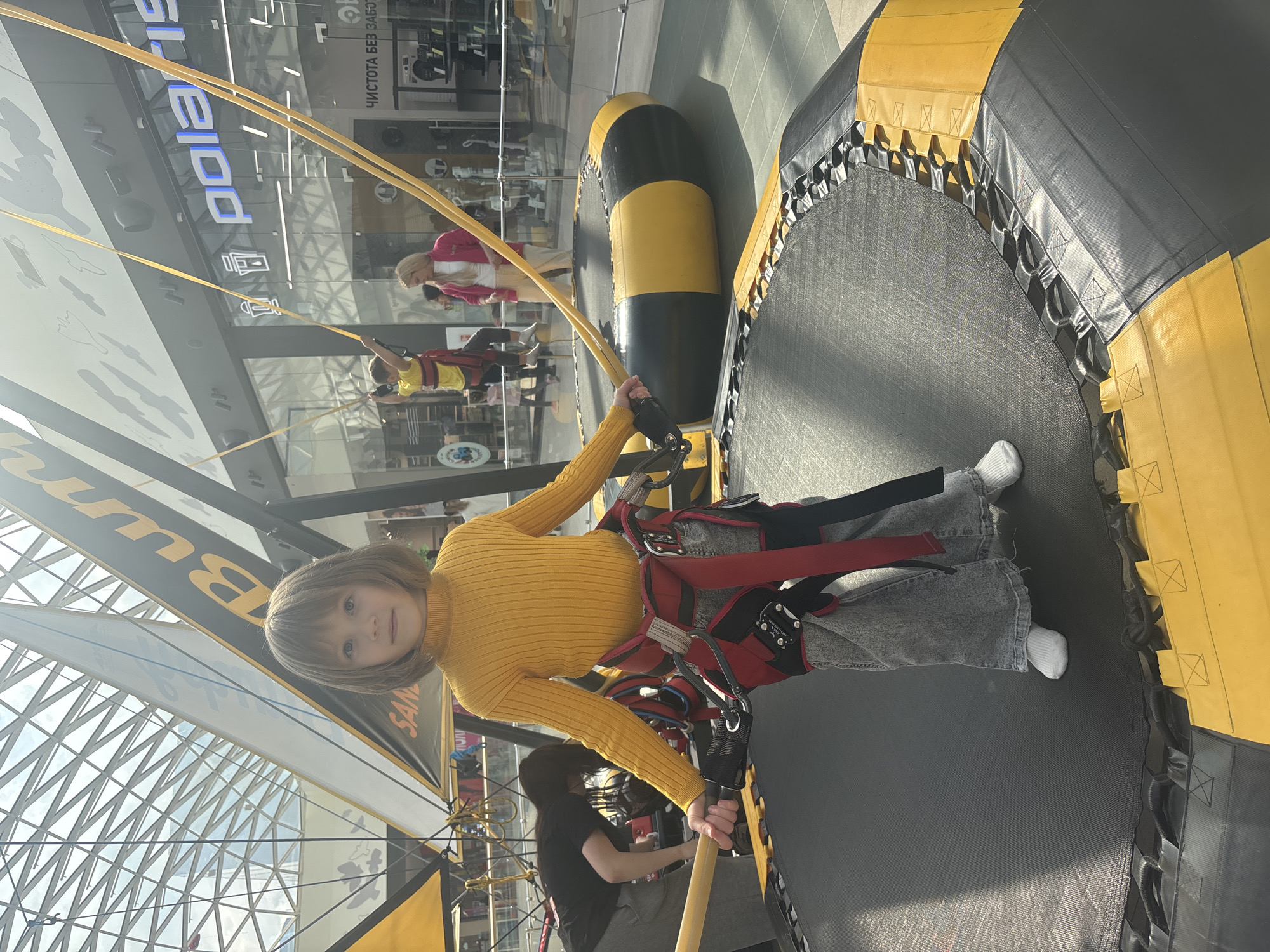 The image captures an indoor bungee trampoline attraction situated within a multi-story shopping mall in Perm, Russia. The location is identified by the prominent "Polaris" retail store sign, which includes the Cyrillic text "ЧИСТОТА БЕЗ ЗАБОТ" (CLEANLINESS WITHOUT WORRIES) on its window, consistent with a Russian-speaking region.
In the foreground, a child, approximately 6-8 years old, stands on a black, circular trampoline with a yellow and black padded perimeter. The child has shoulder-length light brown hair and is looking directly at the camera with a neutral expression. They are dressed in a yellow ribbed turtleneck sweater, wide-leg grey jeans, and white socks. A red and black safety harness is secured around their waist and upper thighs, with black straps connecting to two cylindrical wooden handles held in each hand. Yellow bungee cords extend upwards from the harness attachment points.
In the mid-ground, to the left, a person with dark hair, wearing a black t-shirt and grey trousers, is bent over near an adjacent trampoline, seemingly engaged with equipment. To the right, a second child, dressed in a yellow t-shirt and a red harness, is observed suspended in mid-air, actively utilizing another bungee trampoline setup. Near this second trampoline, a woman with blonde hair, wearing a pink cardigan over a white top and light-colored pants, stands observing.
The background reveals a brightly lit mall environment featuring a large, grid-patterned glass ceiling overhead. Retail store facades, including the aforementioned "Polaris," line the perimeter. Glass railings separate the trampoline area from the main mall walkway. Large, angular yellow and black support structures for the bungee trampolines are visible, one displaying the partial word "Bunge" in yellow lettering. The floor is tiled.