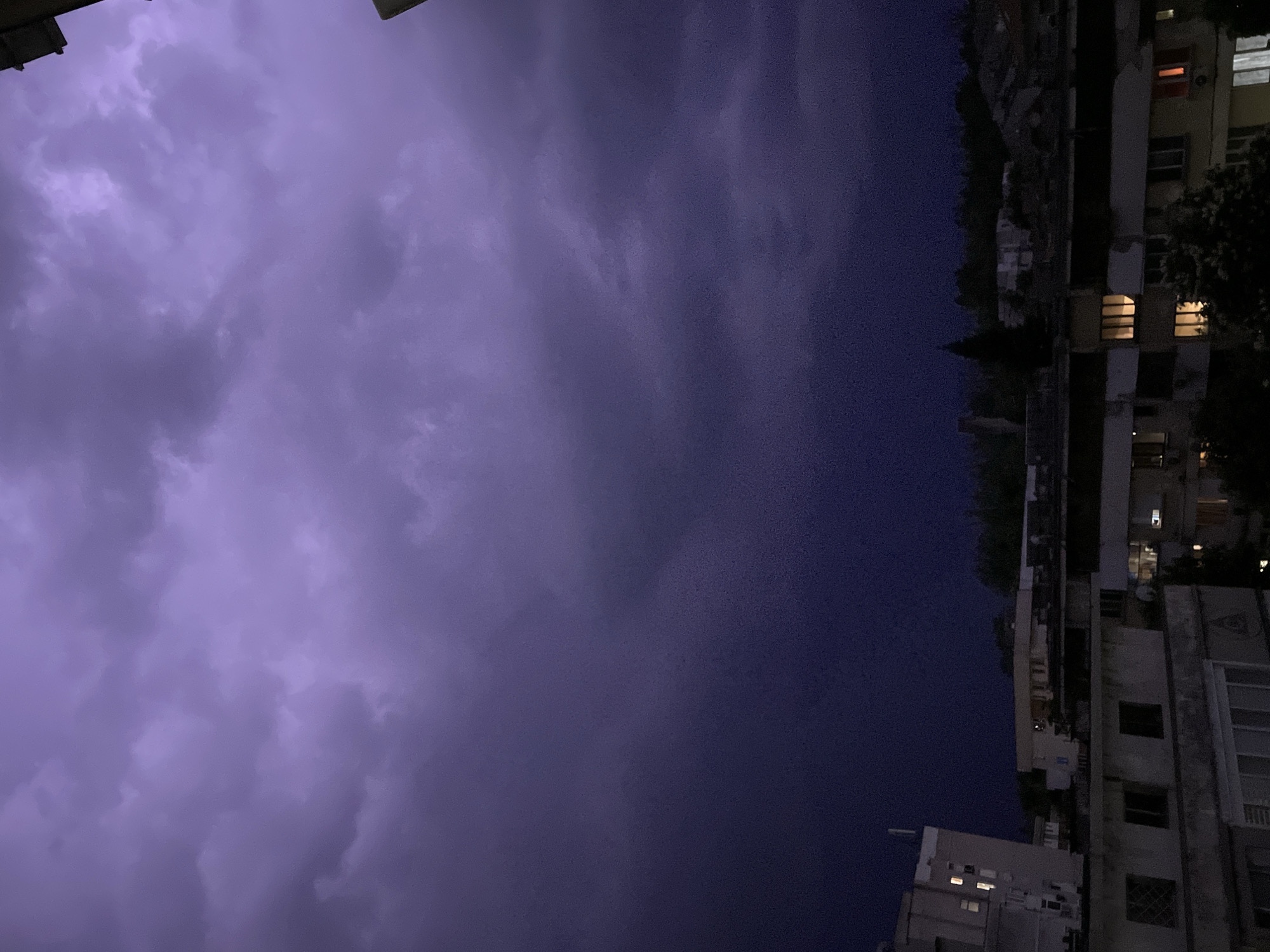 The image shows a night scene in an urban environment. The sky is a dark purple color, indicating an impending storm or nightfall. The right side of the image features a tall building with lit windows, suggesting human habitation. The building's architectural style is not distinctly modern, possibly dating back to the mid-20th century. The photograph is taken from a low angle, looking upwards towards the building and the sky. The precise location is difficult to ascertain without further context, but the presence of a multi-story building suggests a city or town setting.