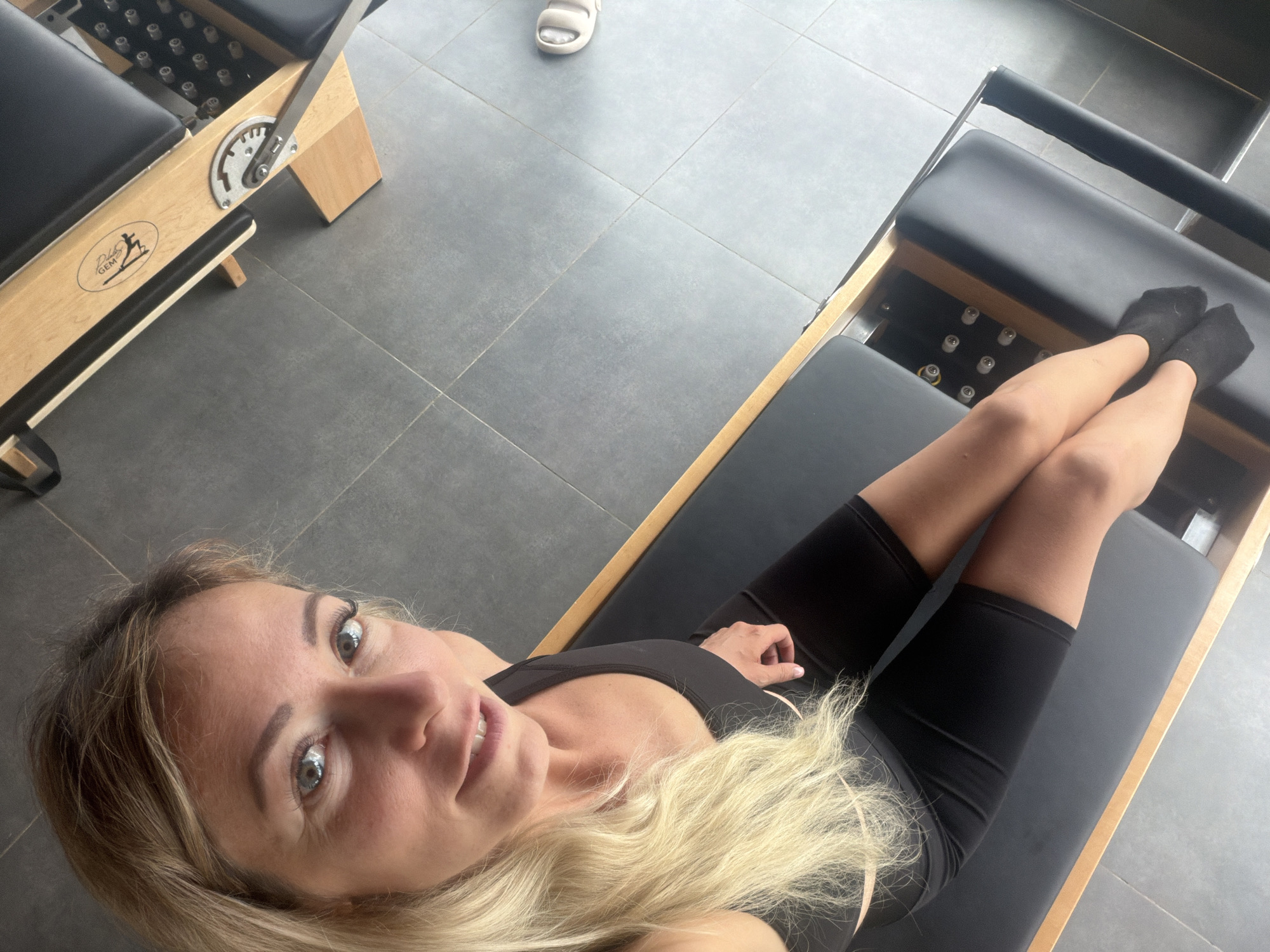 An overhead shot shows a blonde woman in athletic wear sitting on a pilates reformer. She is looking at the camera with a smile. The reformer is made of wood and has black upholstery. The floor is tiled in a gray stone pattern. The scene appears to be an indoor fitness studio. There is no indication of the city of Da Lat, Vietnam in the image.