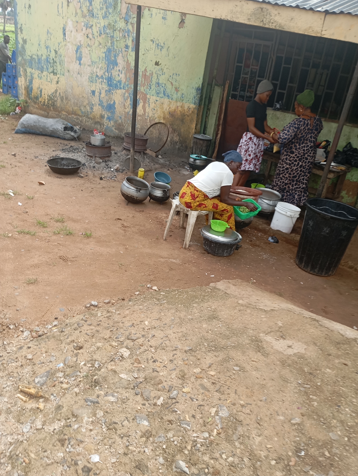 The image depicts an outdoor scene in Shendam, Nigeria, likely a food preparation area or market stall. In the background, a dilapidated building with peeling paint and barred windows stands. To the left, a man is seen stacking blue crates. The foreground is a dirt ground with scattered debris.
Several individuals are engaged in activities. Two women, one wearing a leopard-print dress and a green headscarf, and another in a black t-shirt and a patterned skirt, are standing and interacting near the building entrance. A third woman, seated on a white stool, is wearing a white t-shirt and a yellow and red patterned skirt. She appears to be sorting green vegetables into a green basin.
Various cooking implements and containers are present. Large metal pots and basins are scattered around the area, some appearing to be used for cooking over coals, as evidenced by the ash and embers visible in a large shallow pan. Other items include a large black plastic bin, white buckets, and smaller bowls. A large sack, possibly containing charcoal or grain, rests against the wall. The overall environment suggests a functional, everyday setting focused on food preparation and sale.