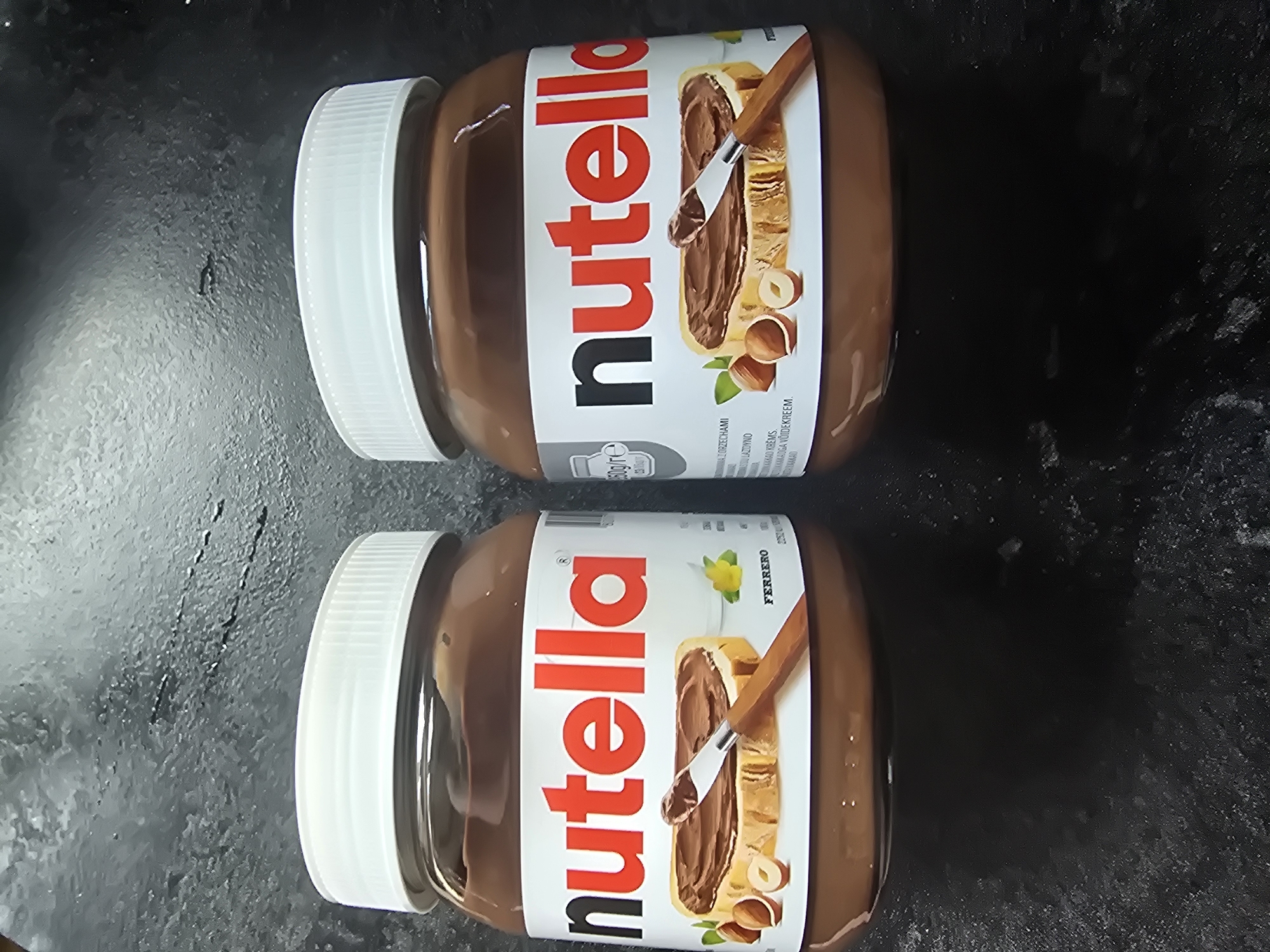 Two jars of Nutella are displayed on a dark, textured surface. The jars are positioned vertically and are oriented so that the labels are facing the viewer. The labels are white with the iconic red "nutella" logo. The upper jar has a partial view of text that reads "FERRERO" and an image of a slice of bread with Nutella spread on it. The lower jar has a similar label with "nutella" prominently displayed. Both jars have white screw-on lids. The surface they are on appears to be a countertop or table with a slightly glossy finish and some subtle texture. The lighting suggests an indoor setting.