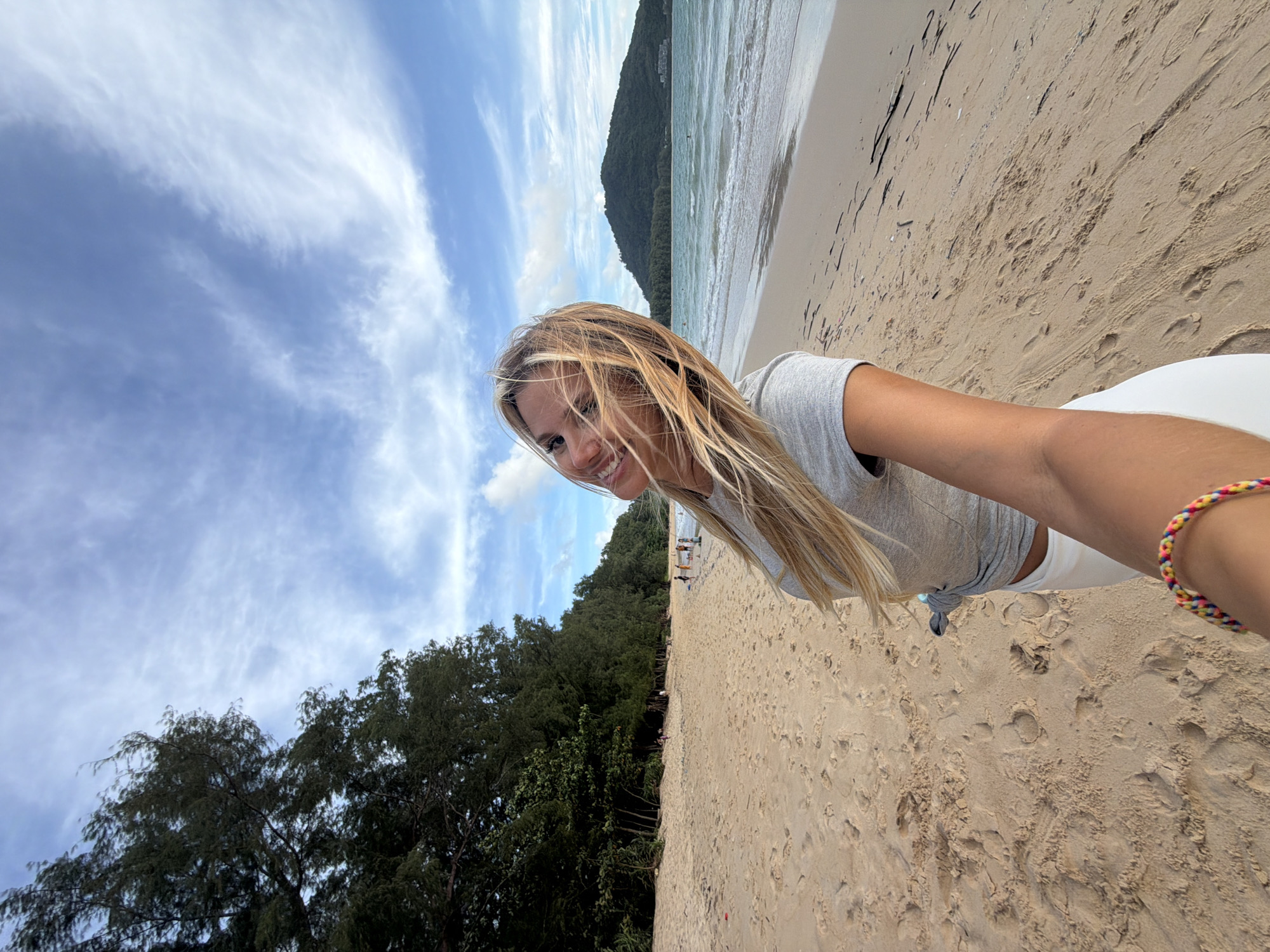 A woman is taking a selfie on the beach in Sakhu, Thailand. She is wearing a grey t-shirt and white pants, and her blonde hair is blowing in the wind. She is smiling and looking at the camera. The beach is sandy, with a line of trees in the background. The ocean is visible in the distance, with a green hill on the horizon. The sky is blue with some white clouds. There are a few other people on the beach, but they are far away.
