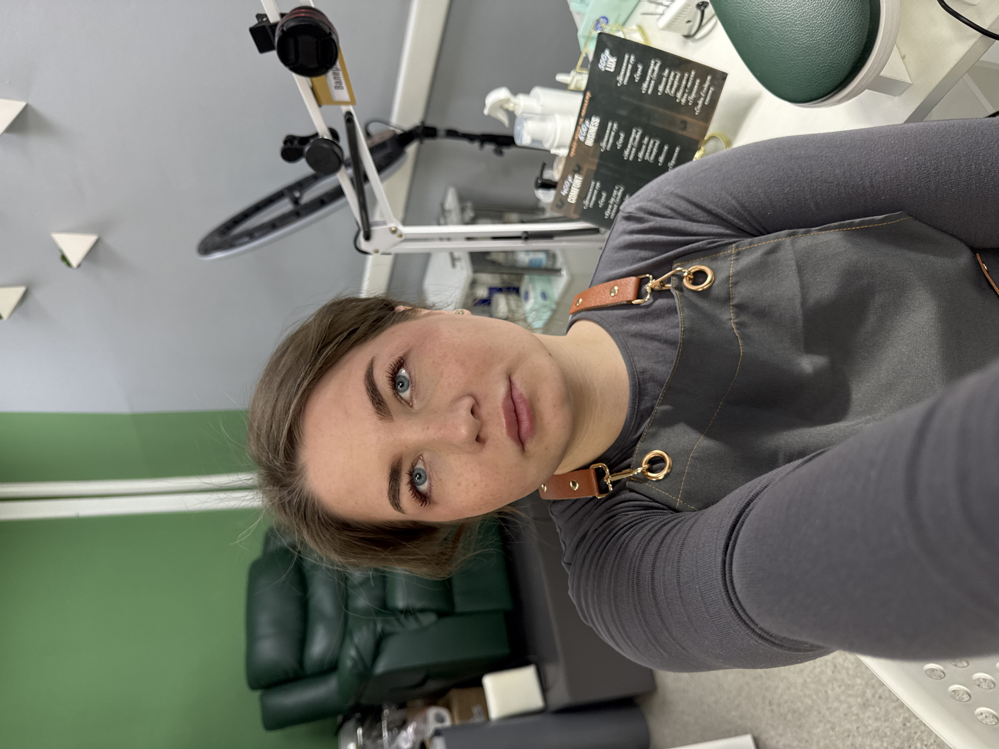 The image is a selfie taken by a fair-skinned woman with freckles and blue eyes. She has light brown hair and is wearing a grey long-sleeved shirt and a grey apron with brown leather straps and gold hardware. The photo is taken indoors, likely in a salon or beauty-related business, as suggested by the equipment and items in the background.
Behind her, there is a green wall and a green leather chair. In the upper part of the image, there is salon equipment, including a ring light and a product menu. The menu lists services in Russian, including "COMFORT," "BUSINESS," and "LUX" options, likely for manicure or pedicure services. The text "Валер" is also visible on a piece of equipment. Given that the services are listed in Russian, the location is likely in Russia. The OCR'd text specifies Russian, and the menu refers to pricing in "400円" which is not Russian. It's unclear as to the city. The environment appears to be a salon setting based on the equipment and furniture.