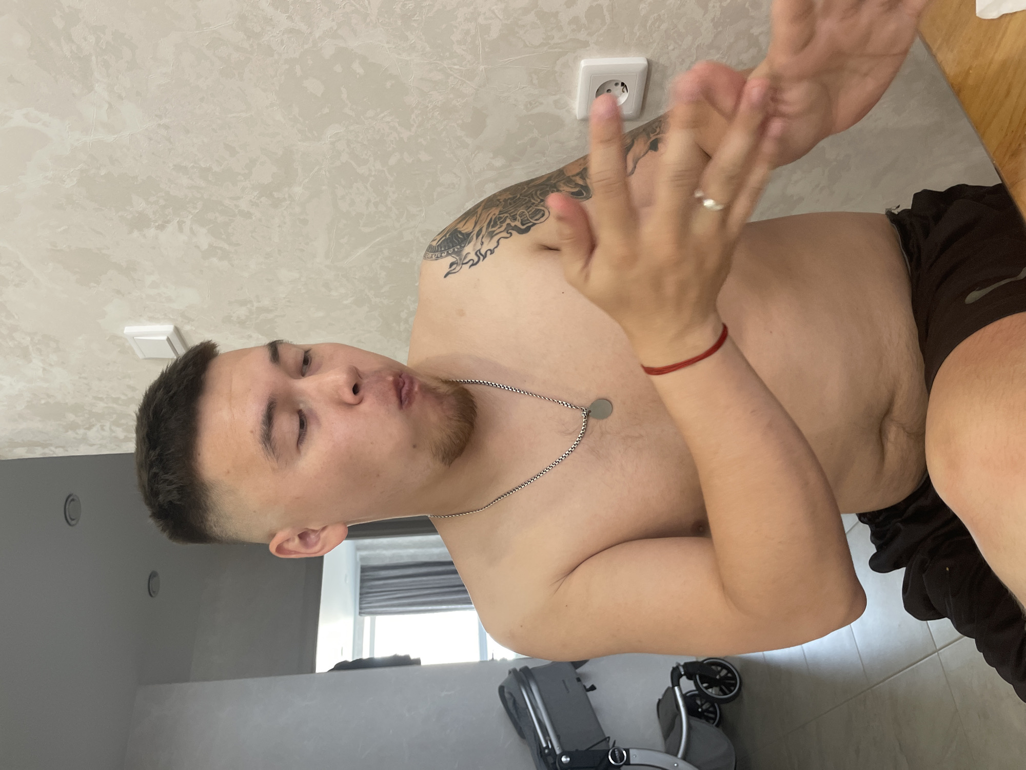 A shirtless man with short, faded brown hair and a goatee is sitting and looking to his left with his mouth open as if eating or speaking. He is wearing a silver chain with a circular pendant, a red bracelet on his left wrist, and dark shorts. He has a tattoo on his left arm. His right hand is raised, and his left hand is gesturing. In the background, a wall with a textured pattern and a white electrical outlet is visible. To the left, a stroller and part of a doorway are visible. The scene appears to be indoors.
