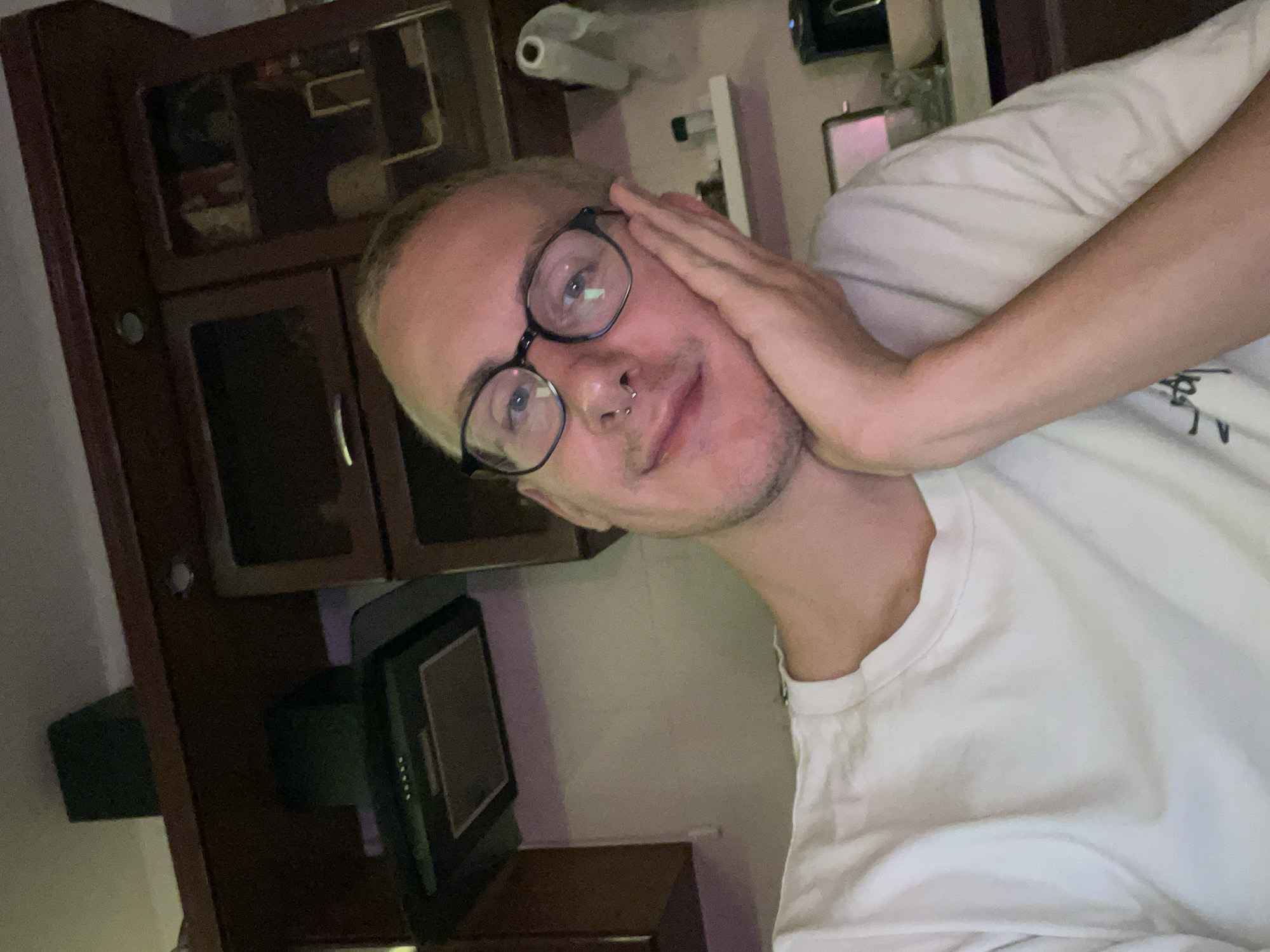 The image shows a person with fair skin, wearing round black glasses and a white t-shirt. They have a nose ring and a hand touching their face. Behind them is a dark brown wooden cabinet with glass doors and a small black television or monitor. The overall lighting is dim, and the setting appears to be indoors. The location in the city of Sestroretsk, Russia, is not discernible from the given image.