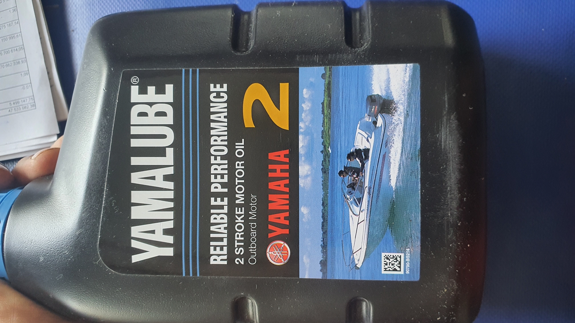 The image shows a black plastic bottle of Yamalube 2 stroke motor oil. The label on the bottle features the Yamalube logo, the words "RELIABLE PERFORMANCE", "2 STROKE MOTOR OIL", and "Outboard Motor". Below this text, the Yamaha logo is prominently displayed in red, followed by a large yellow number "2". The bottom portion of the label contains an image of a white speed boat with two people on a body of water. A QR code and a product number, "90790-BS214", are visible on the lower left of the label, within the boat image. The bottle is held by a person's hand, partially visible at the top of the frame. The background consists of papers with some text, and the surface the bottle is resting on appears to be blue.