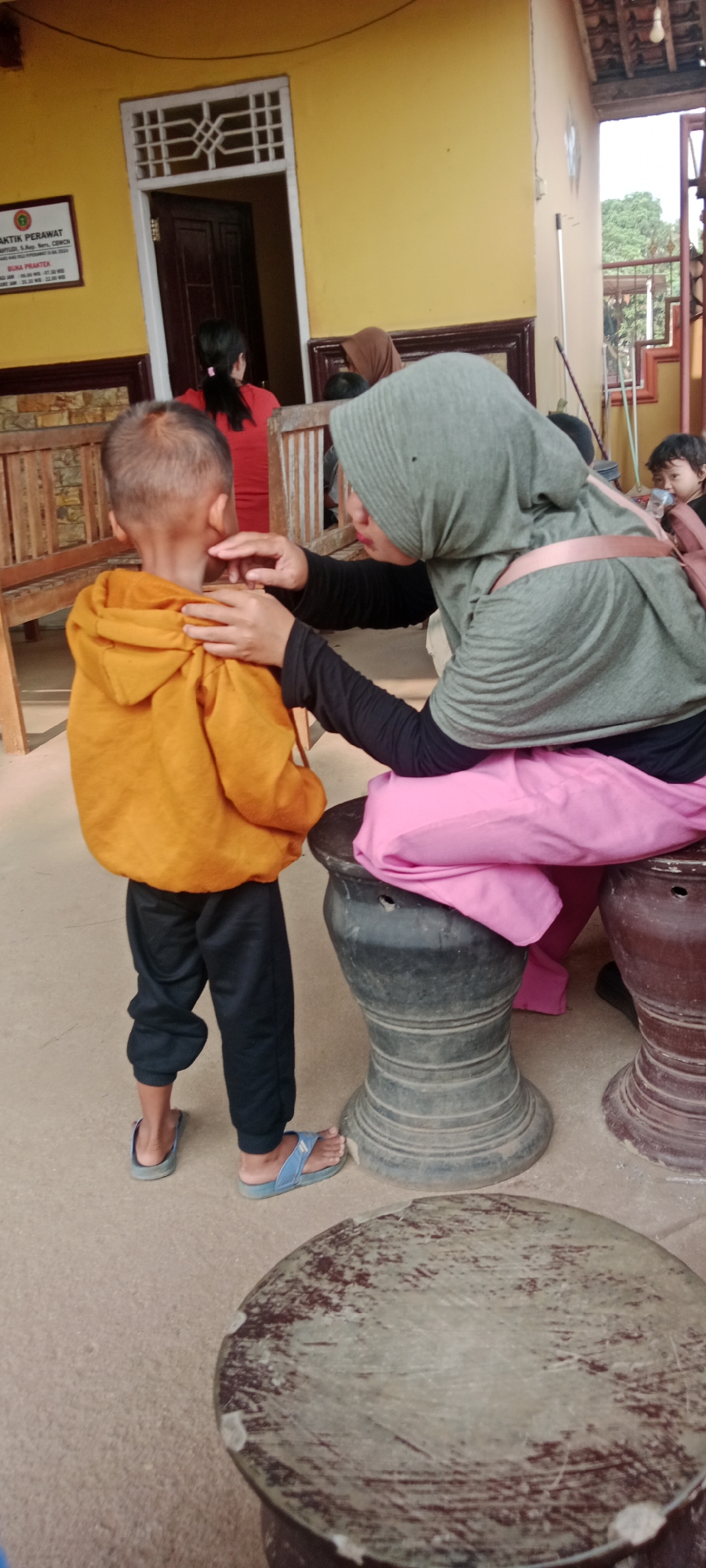 In Sidodadi, Indonesia, a woman wearing a green hijab and black long-sleeved shirt is tending to a young boy in an orange hooded jacket and black pants. The woman is kneeling, and her hands are gently touching the boy's face and neck. The boy is facing away from the camera, towards the woman. Behind them, a yellow wall with a white-framed window and a dark wooden door is visible. To the left, a wooden bench is present, and further in the background, another person in a red shirt is standing. The ground is a light brown, sandy surface. Several large, dark, urn-like objects are placed around the area, some with visible openings. In the far background, a glimpse of trees and sky can be seen, suggesting an outdoor or semi-outdoor setting. The overall scene appears to be a moment of care or examination in a simple, perhaps rural, Indonesian environment.