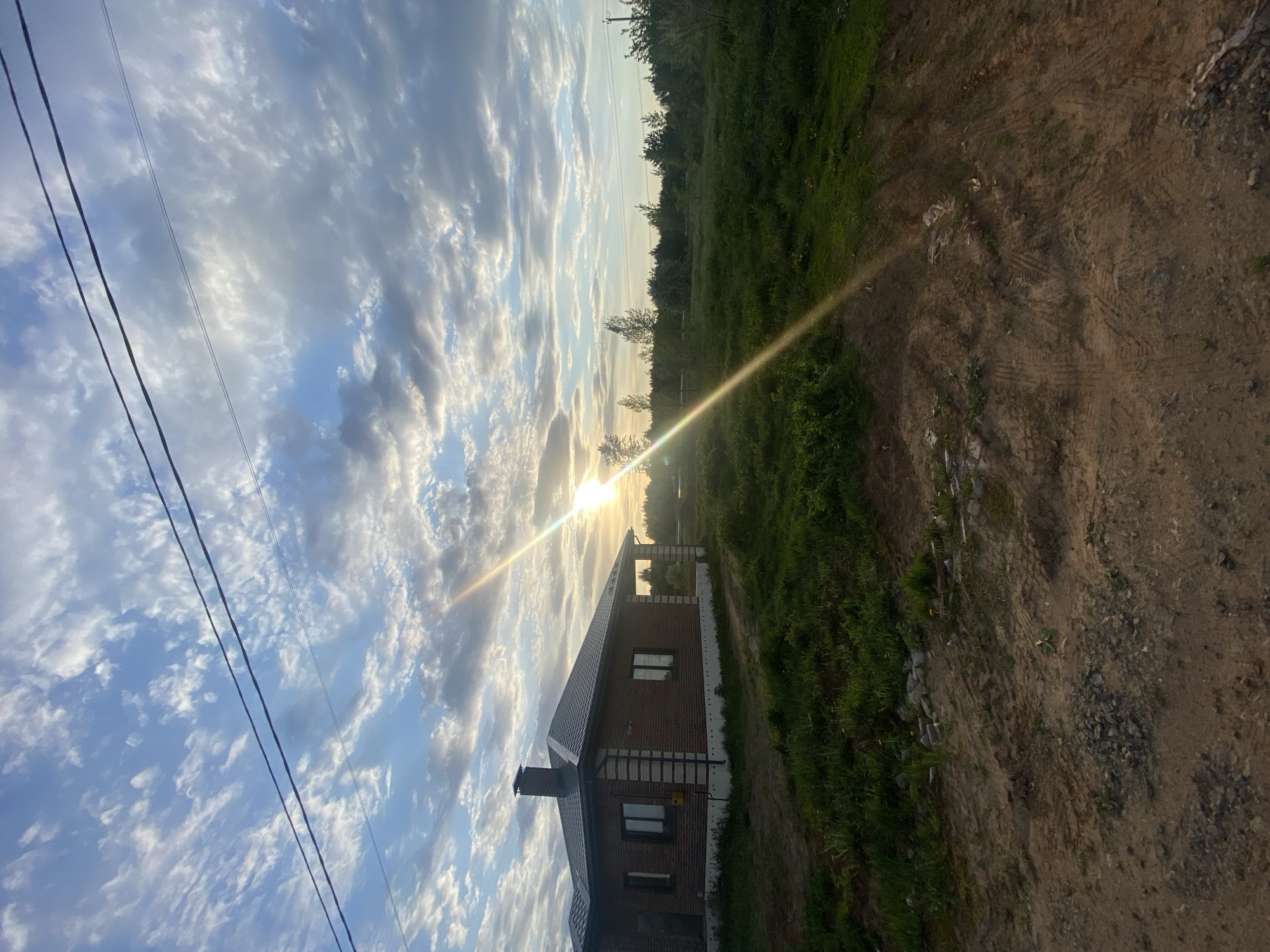 The image depicts a landscape in Prikamskii, Russia, featuring a brick house with a dark roof and chimney. The house is situated beside a grassy area that transitions into a dirt or gravel road. Overhead, there is a sky filled with clouds, with the sun low on the horizon, creating a bright flare in the image. Electrical wires are visible in the upper left corner.