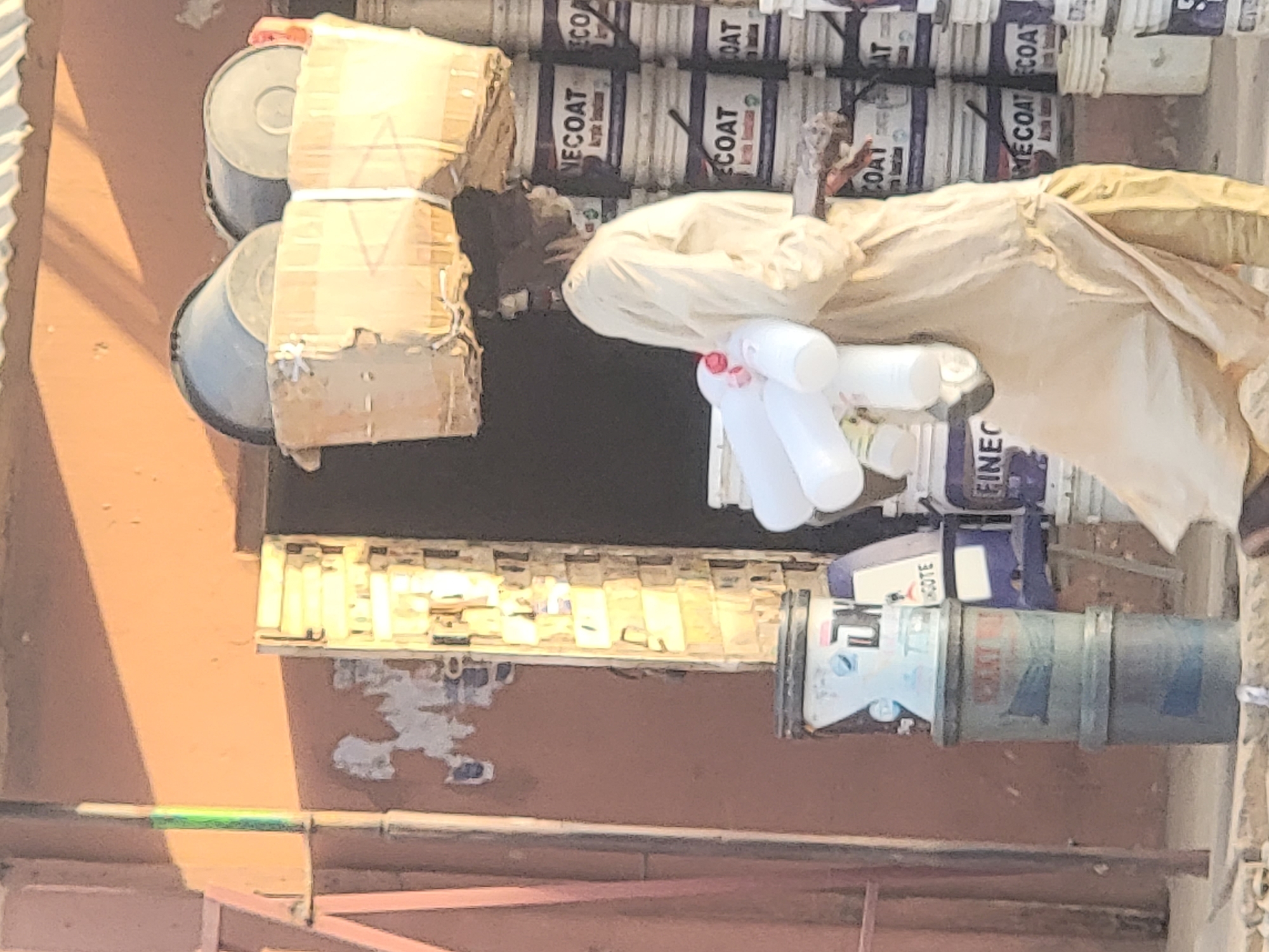 The image shows a person standing among stacks of paint cans in Maiburiji, Nigeria. The individual is wearing light-colored clothing and holding several white plastic bottles. The paint cans are predominantly white with blue and black lettering. Some cans are stacked with cardboard dividers. The background includes a reddish-brown wall with some staining or discoloration. The scene appears to be outdoors, with an overhead structure casting a shadow on the wall. The cans are labeled with "NECOAT".