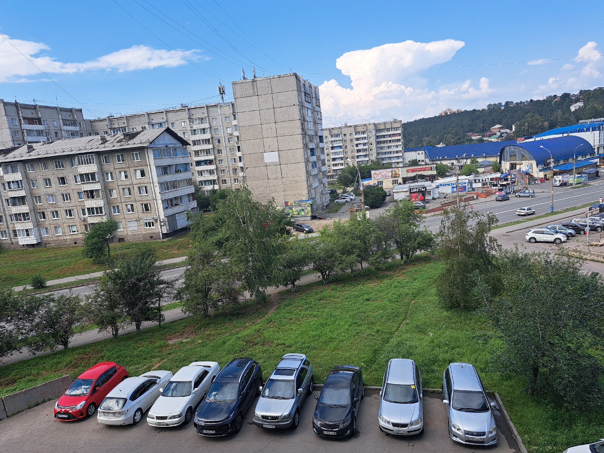 The image captures a view of a city street and residential buildings in Irkutsk, Russia, on a bright, partly cloudy day. In the foreground, a row of parked cars lines a paved area. These include a red hatchback, two white sedans, a dark blue sedan, a silver SUV, a black sedan, and two silver sedans. Behind the cars, a grassy slope leads up to a road with more parked cars and some trees. Further back, several multi-story apartment buildings, typical of Soviet-era architecture, dominate the skyline. These buildings are constructed from grey concrete and feature numerous windows. A large, arched blue-roofed building, likely a market or shopping center, is visible on the right side of the image, with businesses and signs alongside the road. In the background, a densely wooded hill rises, with houses scattered amongst the trees. Power lines crisscross the sky above the buildings and trees. The sky is a vivid blue, punctuated by fluffy white clouds. The scene depicts a common urban landscape in a Russian city, combining residential areas with commercial and natural elements.