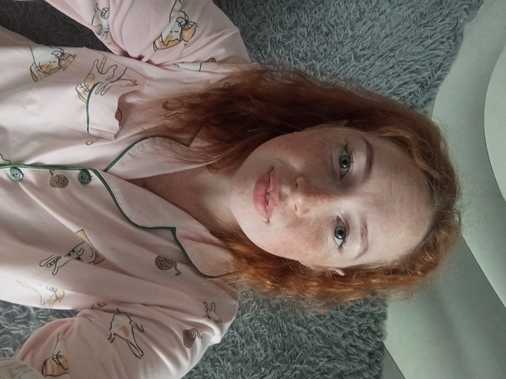 A young woman with red hair and freckles lies on a gray textured surface. She is wearing pink pajamas with a green trim and a pattern of cats and balls of yarn. The scene is indoors. There is no indication of the location being in Yurgamysh, Russia.