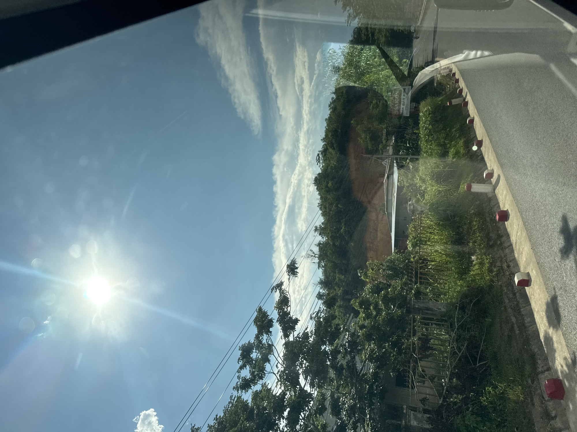 The image depicts a sunlit, blue sky with scattered clouds, as viewed through a vehicle's window. The sun is bright and high in the sky, creating a lens flare effect. Below the sky, lush green trees and vegetation dominate the landscape, lining a road. The road itself appears to be paved, with a concrete curb and red and white bollards. In the middle ground, there are houses and a fence. A sign with Vietnamese text, possibly an advertisement or direction, is visible on the right side of the road. The overall scene suggests a roadside view in a tropical or subtropical region, consistent with Nam Khánh Vĩnh, Vietnam, likely during daytime. There are no people clearly visible in the image.