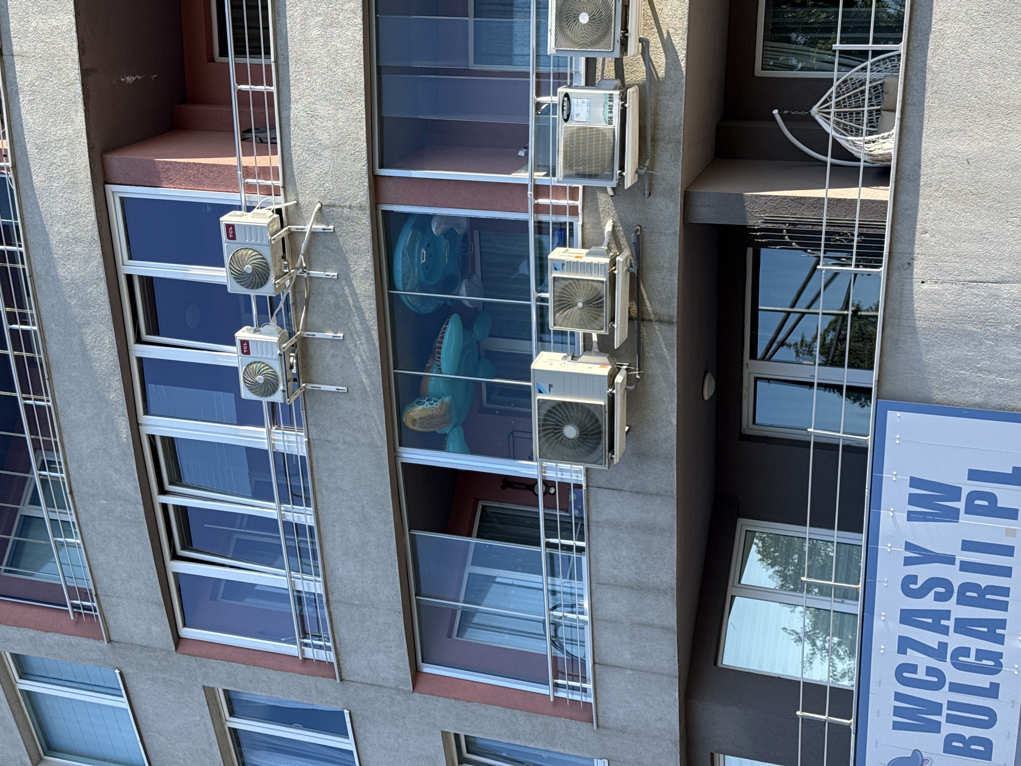 The image shows the facade of a multi-story apartment building in Nesebar, Bulgaria, featuring several balconies and windows. Multiple air conditioning units are visible mounted on the exterior walls, attached to brackets. On one of the balconies, large inflatable toys, resembling sea creatures, are placed inside an enclosed glass-fronted area. Below the balconies, a large blue and white sign with Polish text, reading "WCZASY W BUGARII.PL," which translates to "Holidays in Bulgaria," is displayed. The building has a concrete facade with some sections painted pink. Trees and sky are reflected in the windows.
