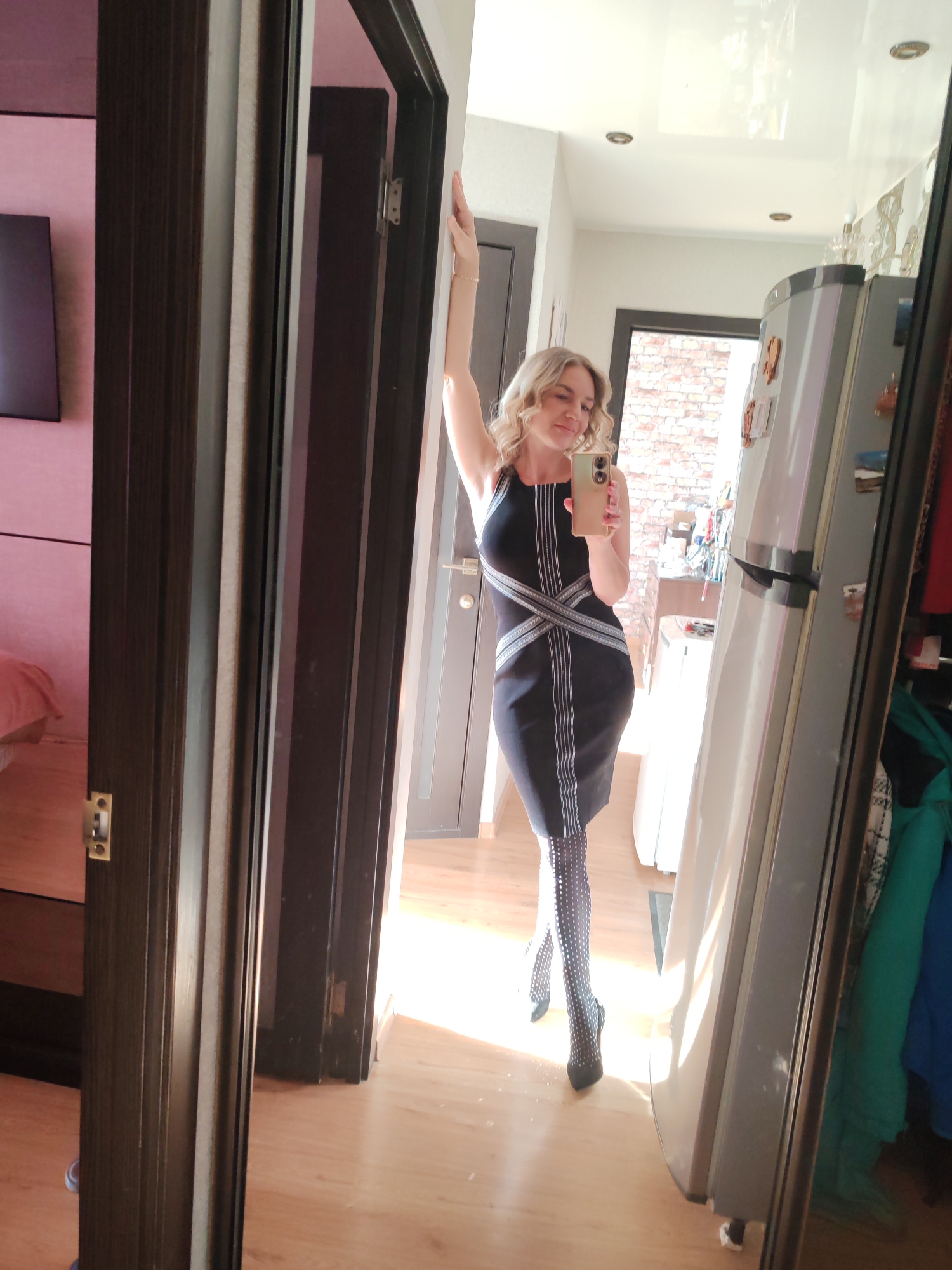 A medium shot shows a woman posing in front of a mirror. She is holding a mobile phone in her right hand and appears to be taking a selfie. The woman has blonde hair and is wearing a black dress with white lines across the torso, along with patterned tights and black heels. She is leaning against a dark doorframe on the left. The reflection in the mirror shows a hallway with a dark door in the background and a silver refrigerator to the right. The floor is made of light wood. The background is illuminated by natural light, and there's a brick wall visible in the distance. There is no information to indicate the location is Petropavlovsk-Kamchatsky, Russia.