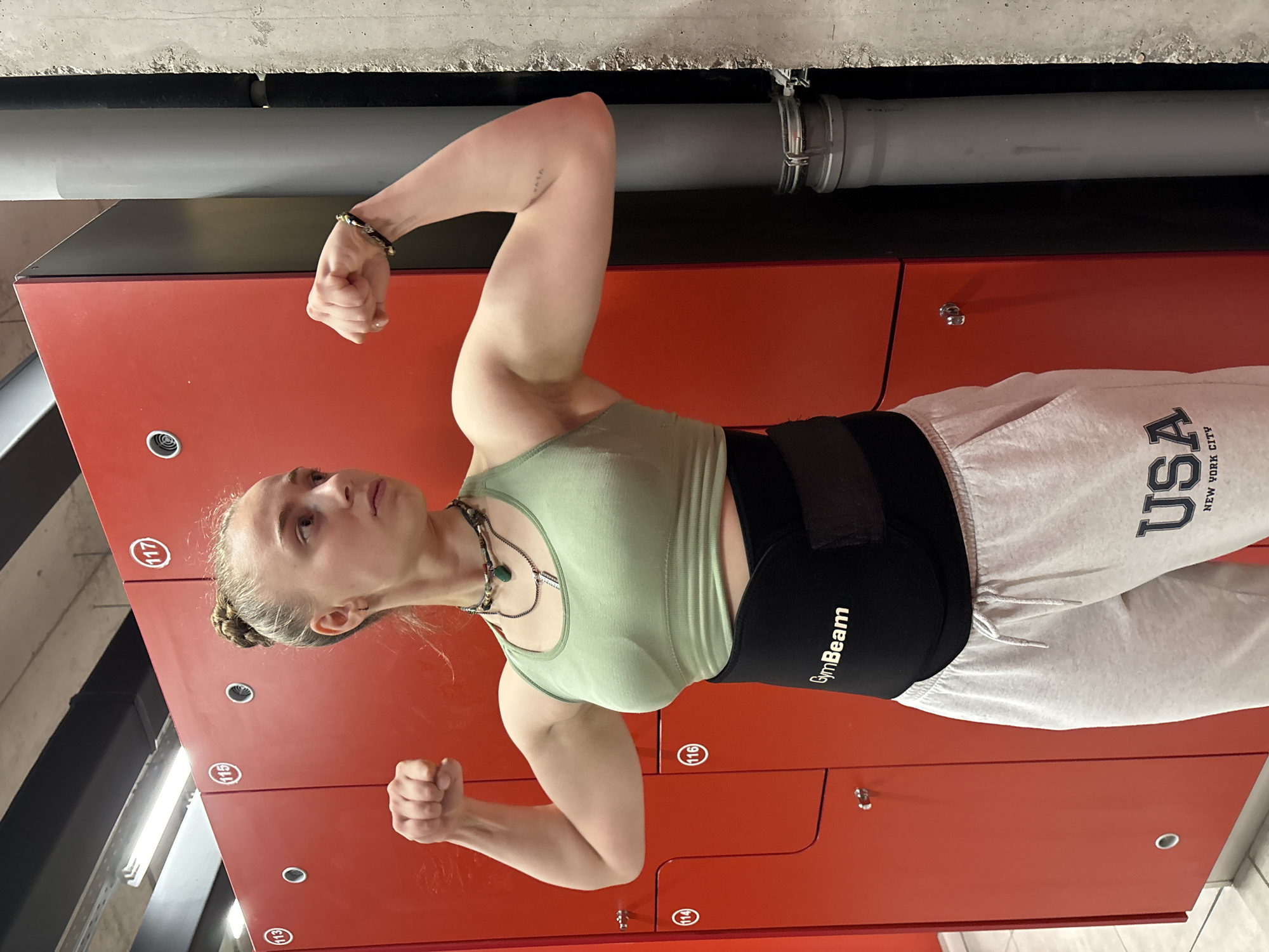 A young adult female is posing in a gym locker room, standing in front of a row of red lockers. She is wearing a light green tank top, grey sweatpants with "USA New York City" printed on the leg, and a black GymBeam branded weightlifting belt. She is flexing both arms, showing off her biceps. The room has a concrete ceiling with exposed piping. The lockers have numbered labels (111-117). The location and city are not identifiable from the image alone.