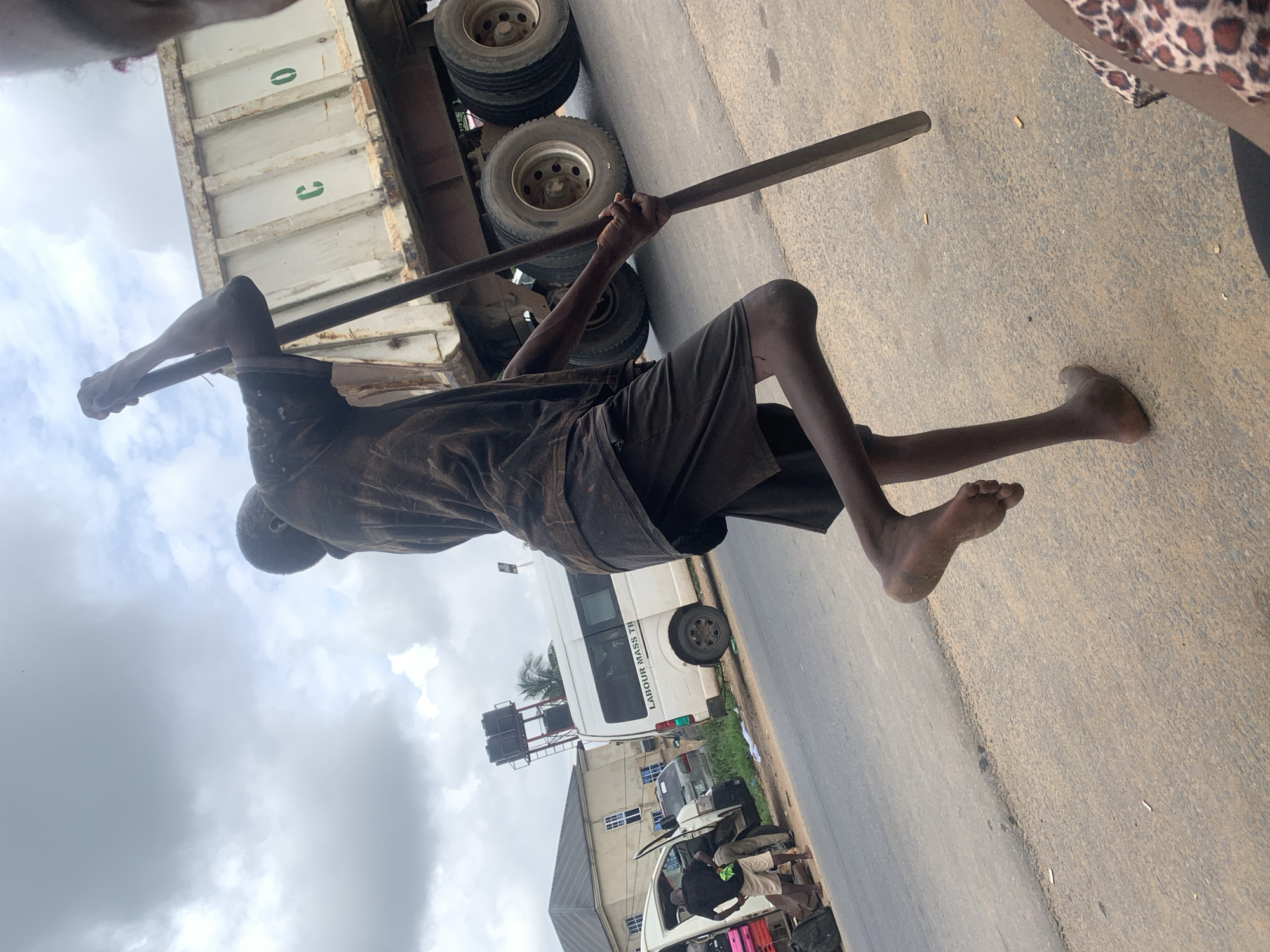 This media file shows a person, likely a young boy, lying down with a long stick across his body. He is wearing dark shorts and a dark t-shirt. His legs are bare and dusty. He appears to be resting or perhaps sleeping on a paved road next to a large truck. The truck has a white cargo bed with green markings that resemble the number "0" and the letter "G". The tires of the truck are visible. In the background, there is a white bus with text that reads "LABOUR MASS TR". There are also buildings and other vehicles, including a white van with its doors open, suggesting a roadside scene. The sky is overcast with clouds, indicating a potentially cloudy or rainy day. The overall setting appears to be Umuahia, Nigeria. The emotion conveyed is one of possible exhaustion or a moment of rest in a busy or working environment.