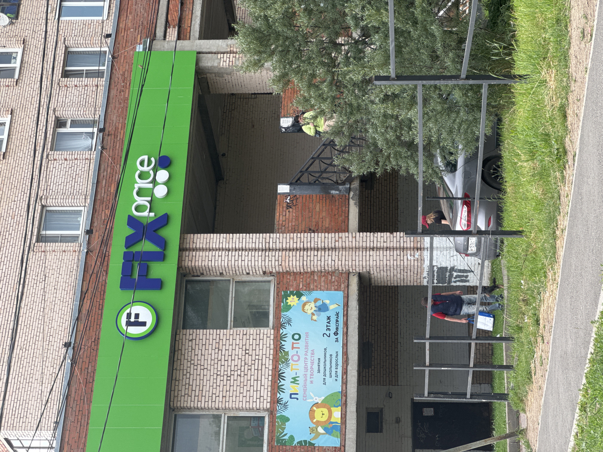 In Kuzmolovskiy, Russia, a building facade displays a bright green sign for "FIX Price" with a circular logo featuring a stylized "F". To the right of the sign, a smaller banner advertises a "Family Center for Development and Creativity" named "LIM-PO-PO", with illustrations of children and animals. The banner also indicates that classes are held on the 2nd floor behind Fix Price.
Several people are visible around the building. A person in a bright yellow jacket appears to be working on the upper part of the building. Near a parked silver car, a woman in a red hat is standing. Further down, a man in a blue vest and red shirt is holding a white bag, possibly near an entrance or walkway. The scene suggests a typical daytime activity in an urban or suburban setting, with clear weather. Power lines run vertically along the brick building. The overall impression is of a commercial area with community services.