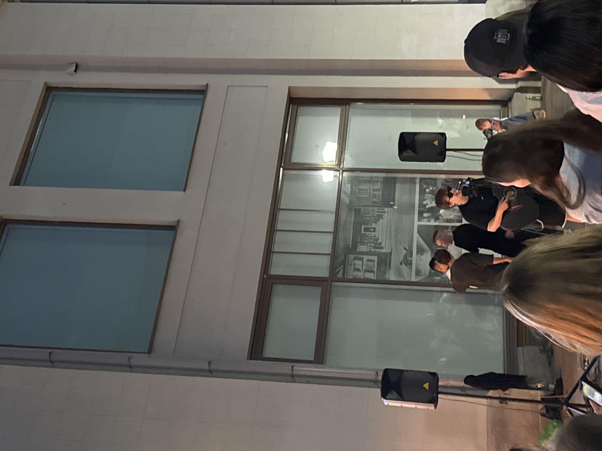 The image depicts an outdoor performance in front of a modern building with large windows. A young man in a black t-shirt and sunglasses is playing an acoustic guitar and singing into a microphone, suggesting a musical performance. He is accompanied by other individuals, possibly bandmates or attendees, some of whom are gathered around him. Two black speakers are visible, indicating amplification for the performance. The background features a building facade with a large mural or decorative window covering depicting a street scene. Several people are visible in the foreground and mid-ground, watching the performance. The scene appears to be taking place in the evening or at dusk, with artificial lighting illuminating the area. The architecture and style of the building, combined with the presence of a specific mural, suggest the location is on **Vaynera Street in Yekaterinburg, Russia**, known for its pedestrian nature and street art.