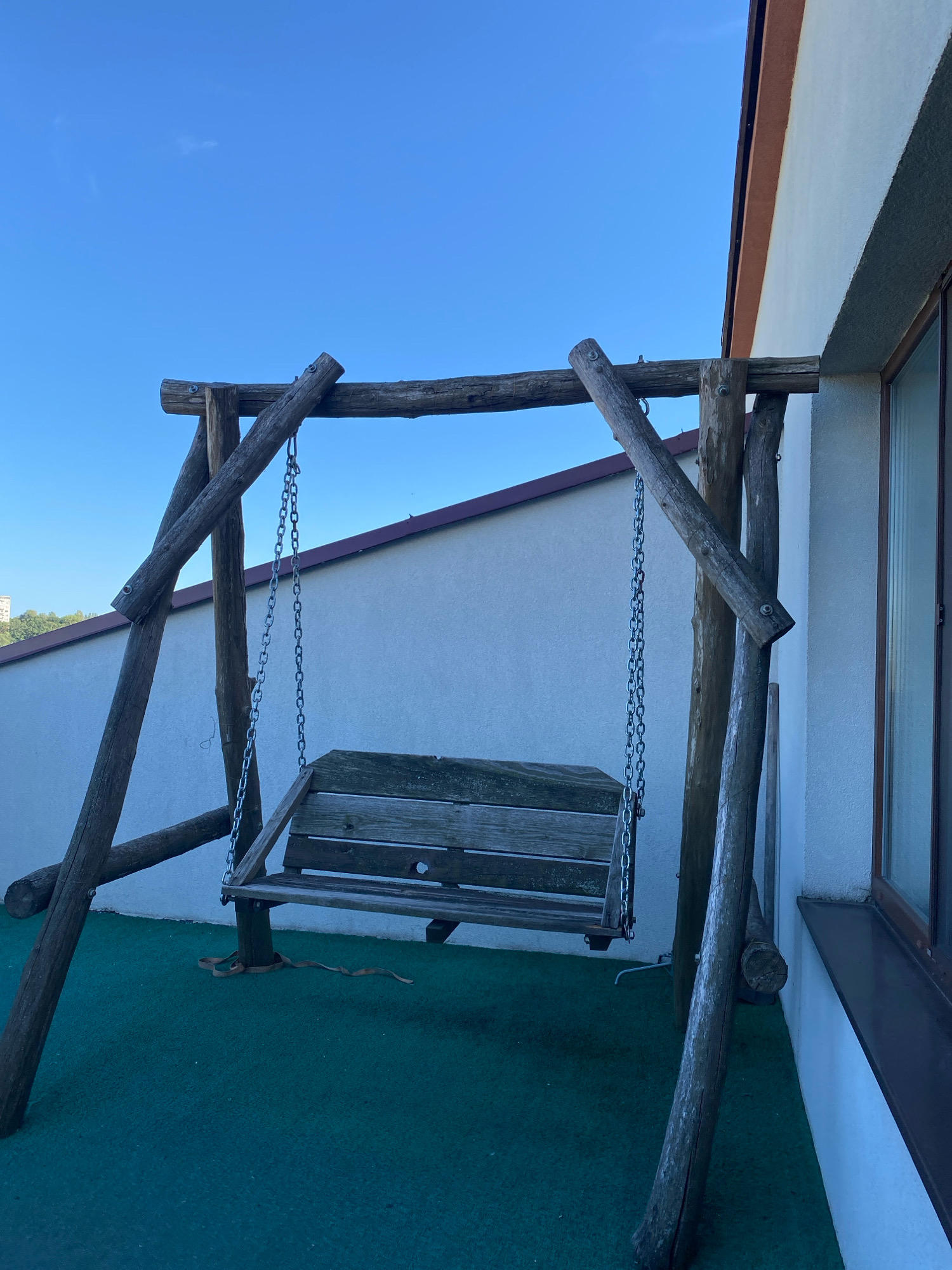 The image shows a rustic wooden swing set on a green surface, possibly artificial turf, in an outdoor urban setting. The swing itself is made of weathered wooden planks and hangs from metal chains. The swing set is constructed from rough-hewn wooden logs, giving it a natural, handmade appearance.
In the background, a light-colored building with a brown trim along the roofline is visible, along with a window with a dark frame. The sky is a clear, bright blue. The setting appears to be a balcony, patio, or rooftop, given the proximity to the building and the elevated perspective.
While the exact location in Chișinău, Moldova cannot be determined from the image alone, the architectural style of the building and the presence of what appears to be a residential or commercial structure in the distant background (visible on the left edge of the image) are consistent with urban environments in the city. There are no people or animals present in the image. The scene is static and depicts a quiet, outdoor space.