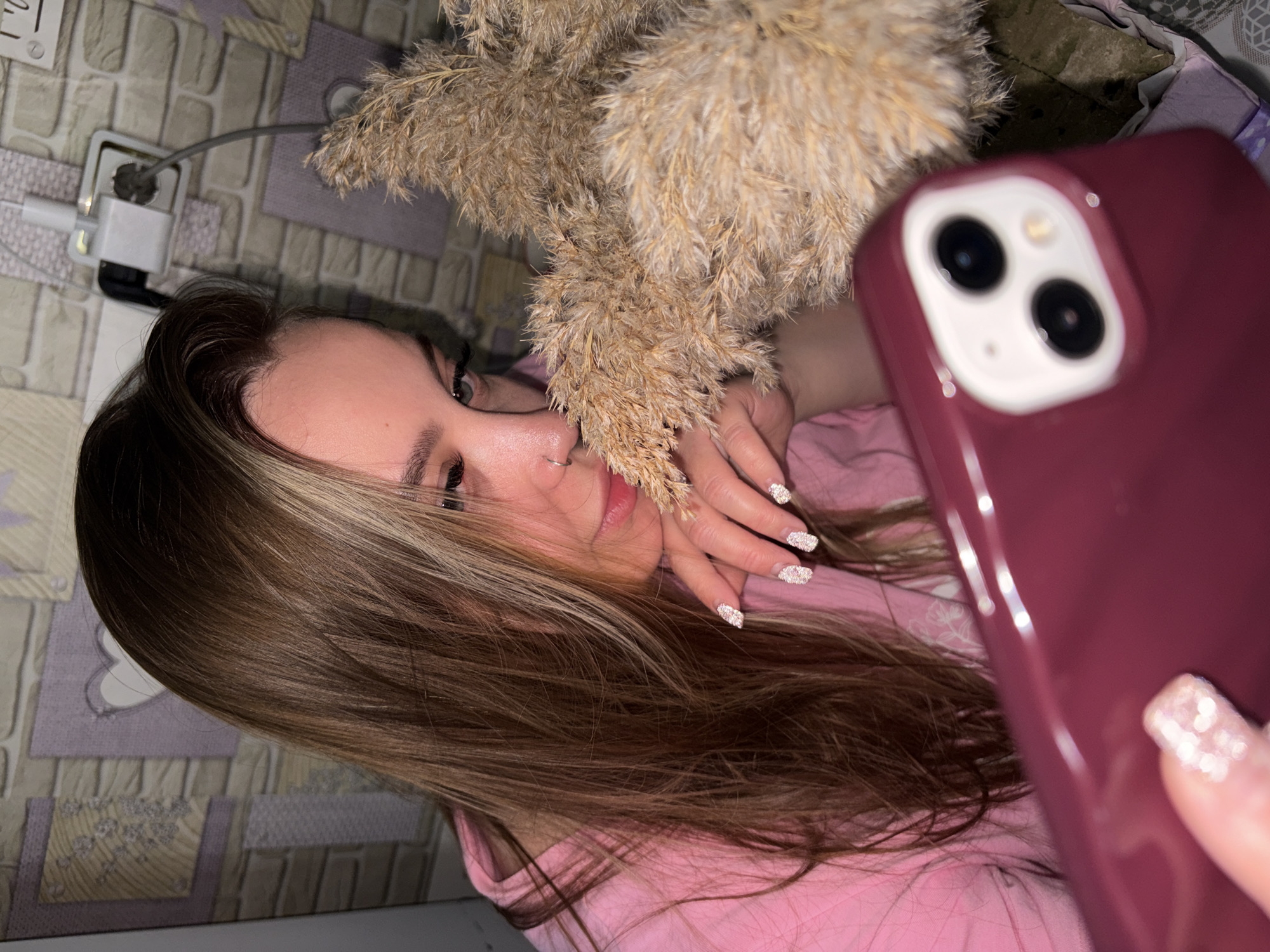 A young woman with long, brown hair with blonde highlights takes a selfie with a red iPhone. She is wearing a pink shirt and has a silver nose ring. Her nails are long and painted with white glitter. She is holding a bouquet of dried pampas grass in front of her face. The background is a textured wall with an electrical outlet and cord visible in the upper left corner. The scene suggests an indoor setting, possibly a bedroom or living room. There is no indication of the city of Port-Katon, Russia, within the image.