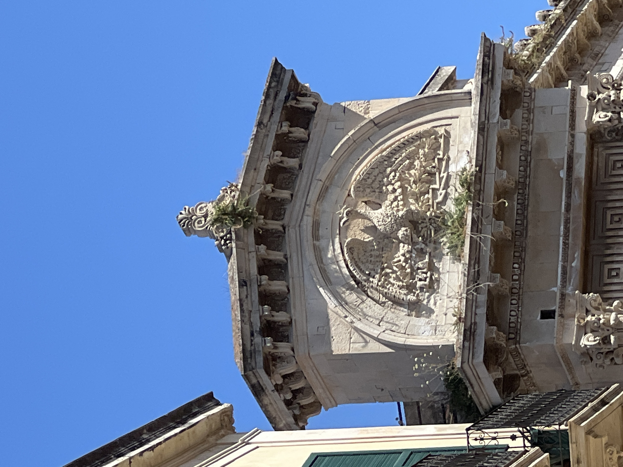 An ornate stone relief sculpture of a bird, possibly a phoenix, is prominently displayed on a building in Syracuse, Italy. The sculpture is housed within an arched recess and is surrounded by intricate carvings of what appears to be fruit or foliage. The bird is depicted with its wings spread, facing left, and its detailed feathers are visible. The relief is weathered and shows signs of age, with small plants growing in crevices. Above the arch, the building features classical architectural elements, including corbels and decorative moldings. To the left of the main structure, a portion of an adjacent building is visible, with a balcony featuring an iron railing and a green shutter. The sky is clear and blue, indicating a bright, sunny day. The image captures a detailed view of architectural ornamentation against a simple background.
