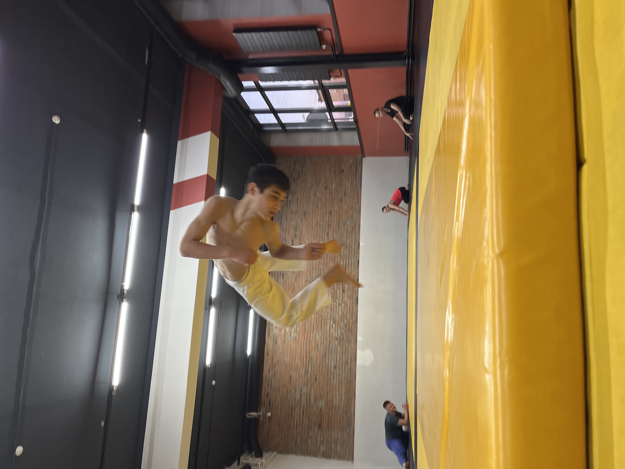 The image depicts a shirtless male wearing white pants mid-air, appearing to perform a martial arts kick. He is holding an object, possibly a piece of chalk or padding, in his hand. The environment appears to be an indoor parkour or climbing gym, evidenced by the yellow padded wall on the right, the exposed brick wall in the background, and other individuals climbing or observing in the distance. The gym has a ceiling with multiple window panes that allow light to flood the area.
Given the information, the location is Krasnoyarsk, Russia.