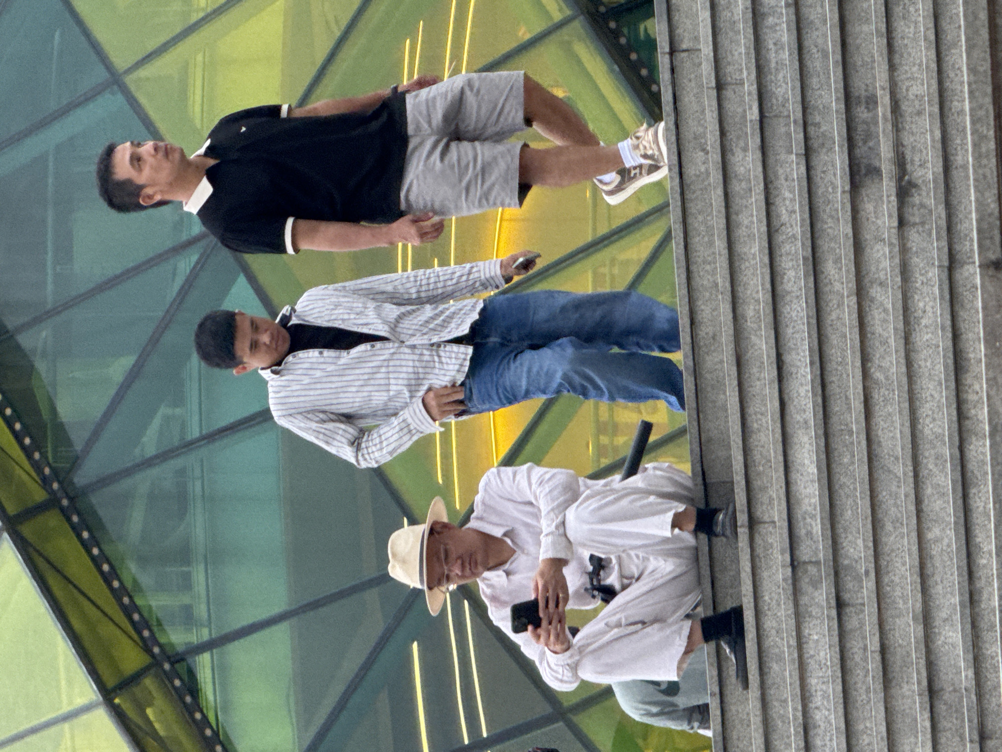 Three men are standing on a set of outdoor stairs in front of a modern building with a geometric facade. The man on the far left is wearing a light-colored hat, glasses, a light-colored shirt, and light-colored pants, and is sitting on the stairs, looking at his phone. The man in the middle is wearing a striped shirt and blue jeans, and is walking down the stairs. The man on the right is wearing a black polo shirt and grey shorts, and is walking down the stairs. The building behind them has a facade made of green and yellow geometric shapes. This scene is observed to be in Da Lat, Vietnam.