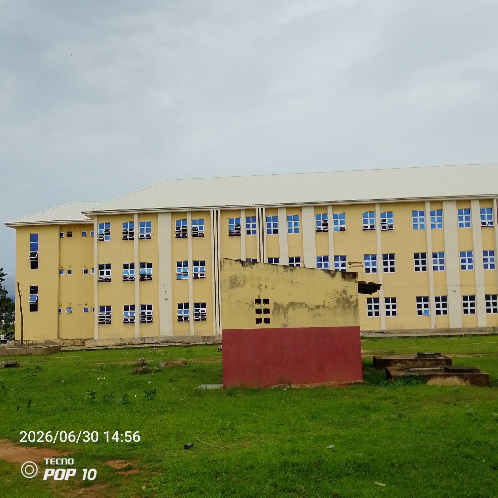 A long, pale yellow, multi-story building with a white roof stands under an overcast sky in Jalingo, Nigeria. The building has numerous windows with blue panes, many of which are open. In the foreground, a patch of green grass is dotted with small plants and some litter. To the right of the main building, a smaller, dilapidated yellow and red structure is partially visible. The timestamp "2026/06/30 14:56" and "TECNO POP 10" are superimposed on the lower-left portion of the image.