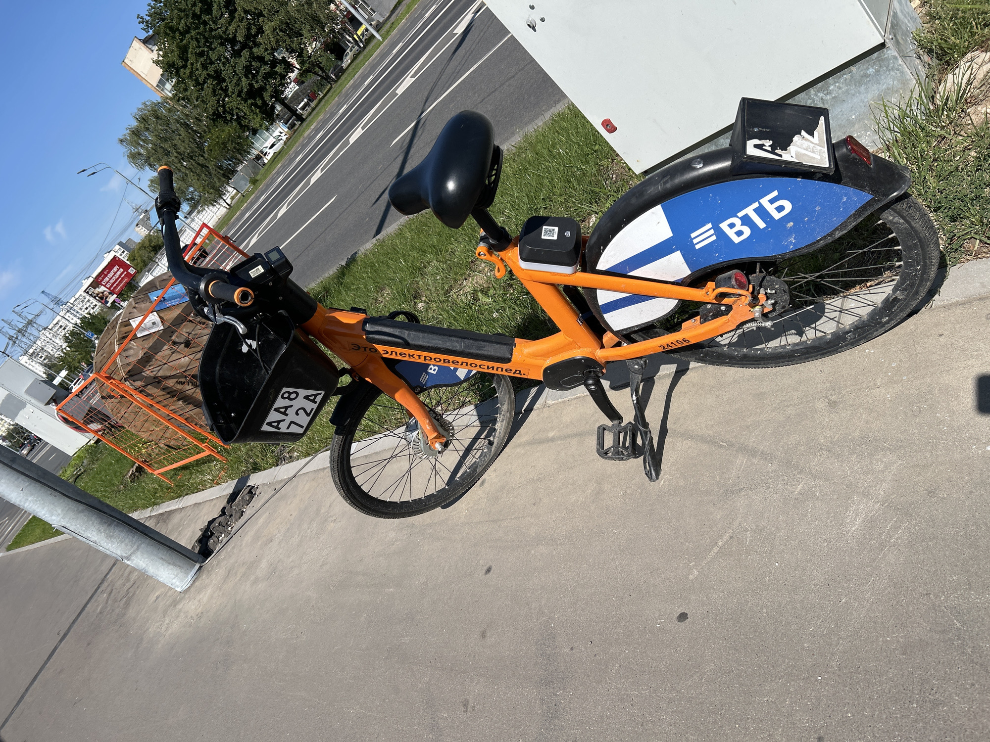 An orange public-use bicycle, identified by the license plate AA8 72A and branding "ЭТО ЭЛЕКТРОВОЗЯЩИЙ", is parked on a concrete sidewalk next to a grassy area and a road in Moscow, Russia. The bicycle has a black basket in the front, a black seat, and a QR code for unlocking. The rear fender is blue and white with the Cyrillic abbreviation "ВТБ". In the background, there are power lines, buildings, trees, and traffic. A construction barrier with orange netting is visible on the right side of the image. The sky is blue and clear.