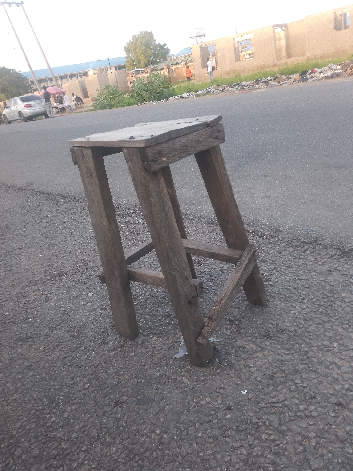 A low-angle, medium shot captures a worn wooden stool placed on an asphalt road. The stool, with its rough-hewn planks and weathered appearance, sits at an angle, with its legs slightly splayed. In the background, a silver sedan is parked on the side of the road, with several people gathered nearby. Beyond the car and people, construction is underway on several buildings, some appearing incomplete with exposed brickwork. Vegetation, including trees and bushes, lines the edge of the road. The sky above is overcast, casting a diffused light on the scene. The overall impression is of a casual, everyday setting in an urban environment, possibly in a developing area.