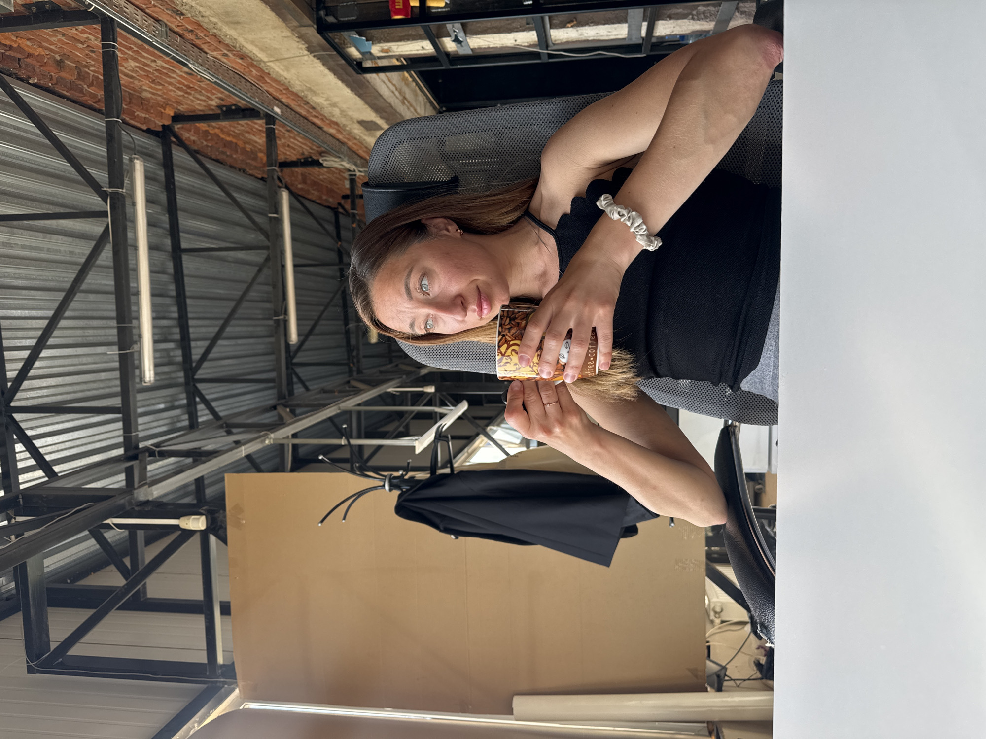 A woman is sitting in an office chair, holding a coffee cup. She has long brown hair and green eyes. She is wearing a black tank top and a white scrunchie on her wrist. The background includes a metal ceiling with lights, a brick wall, a black umbrella, and a cardboard backdrop. It cannot be confidently confirmed this photo was taken in Chaikovsky, Russia.