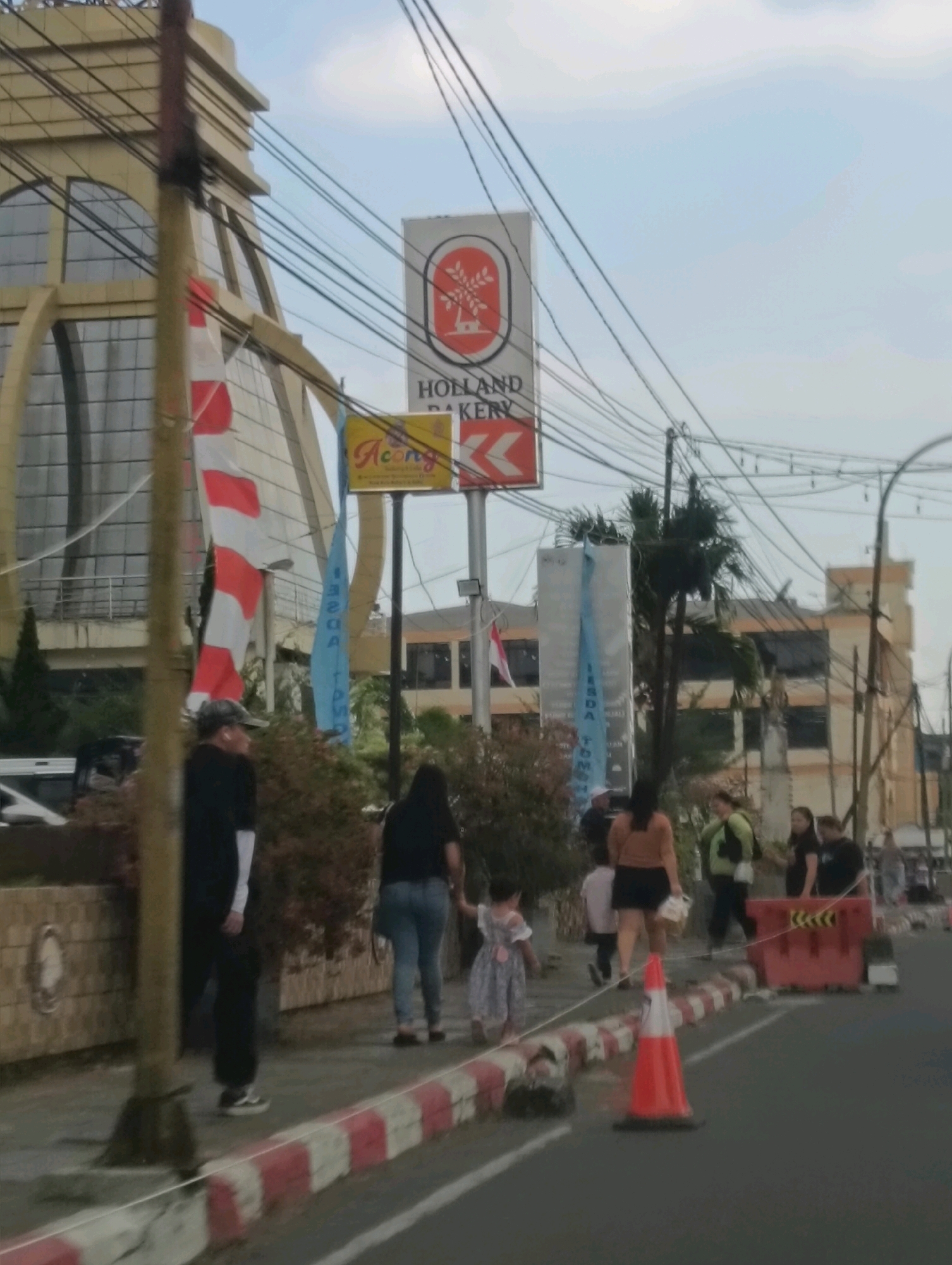 The image depicts a street scene in Tomohon, Indonesia, featuring the Holland Bakery and a building with a distinctive arched facade. Several individuals are visible on the sidewalk and near a red traffic barrier. In the foreground, a traffic cone is positioned on the road. Overhead, a dense network of power lines crisscrosses the sky. The scene appears to be in an urban setting, likely during daylight hours.