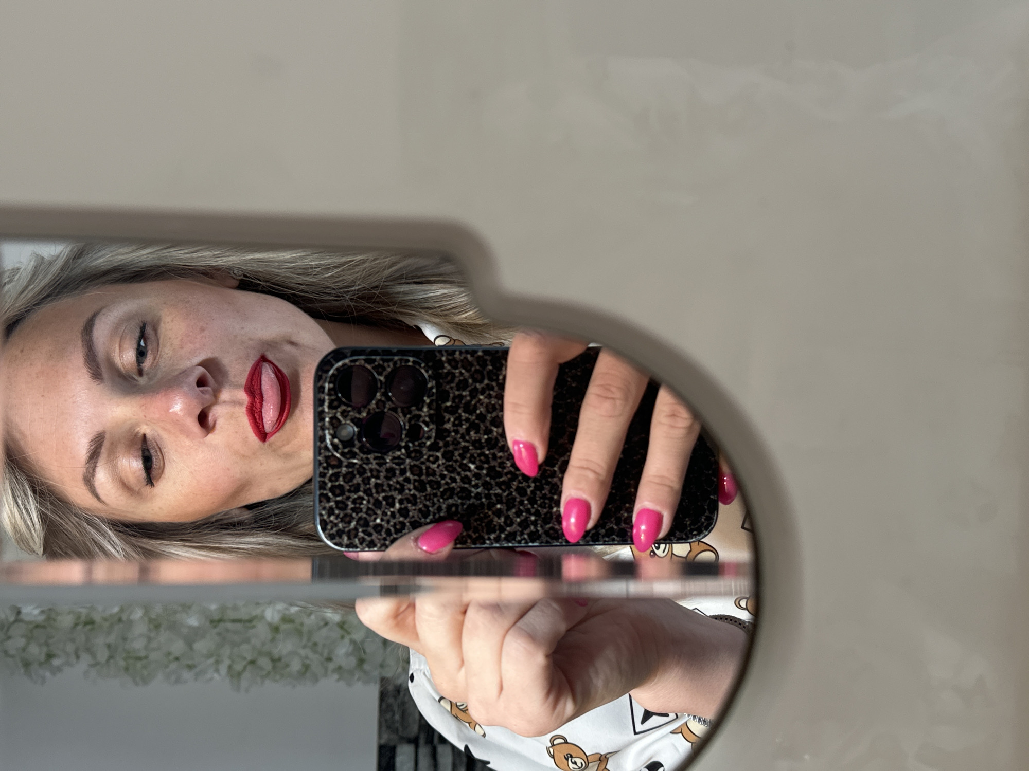 The image shows a woman taking a selfie in a mirror. The reflection reveals her face with red lipstick and a dark-patterned phone held in front of her. She is wearing a white shirt with a teddy bear pattern. The surroundings of the mirror are visible, including a white floral arrangement and a neutral-colored wall. The location is unknown, and there is no information to associate the image with Sesto Campano, Italy.