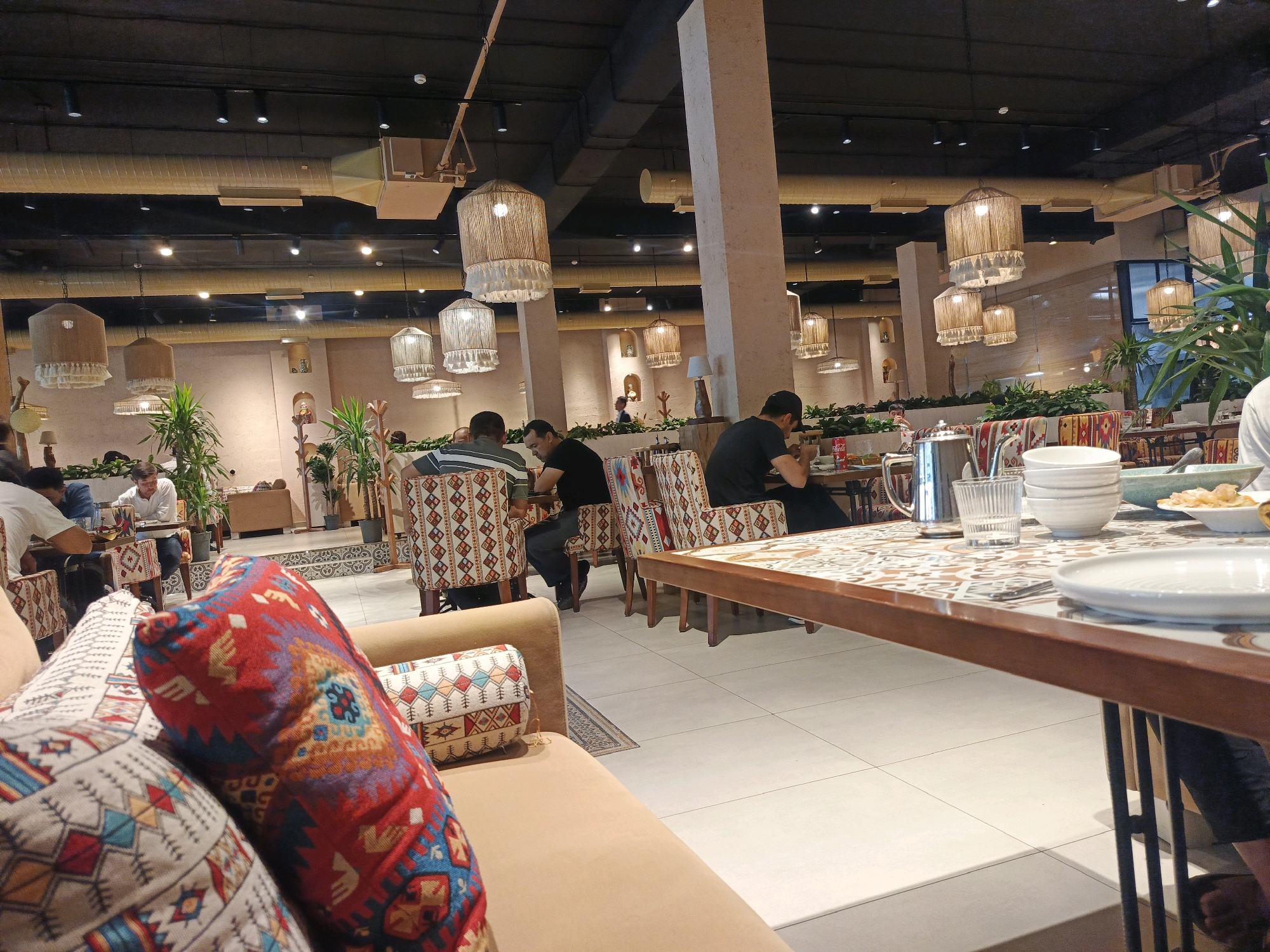 The image depicts the interior of a restaurant in Tashkent, Uzbekistan, likely a modern establishment with a cultural aesthetic. The scene is illuminated by ambient lighting from numerous ceiling fixtures and decorative lamps with woven shades. In the foreground, a plush sofa with vibrant, patterned throw pillows suggests a comfortable seating area.
The middle ground of the image focuses on a dining area with tables and chairs. Several individuals are seated at tables, engaged in dining or conversation. The chairs are upholstered with fabric featuring intricate geometric patterns, reflecting a design influenced by local traditions. On one of the tables, a metallic teapot, glassware, and a stack of bowls are visible, indicating a meal is in progress. The flooring is tiled with a light-colored, large-format tile.
In the background, the restaurant's architecture is apparent, with exposed ceiling ducts and structural elements. Potted plants are strategically placed throughout the space, adding a touch of greenery. The overall ambiance suggests a contemporary dining experience that incorporates elements of Uzbek cultural design.