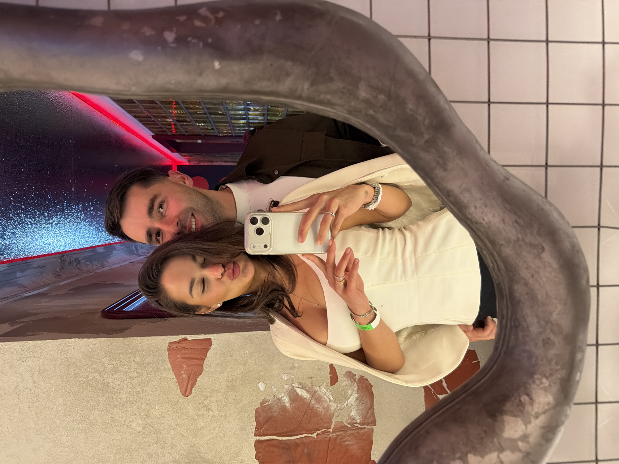 A couple takes a mirror selfie in an indoor setting, likely in Krasnoyarsk, Russia. The image, captured through a distorted mirror, shows a man and a woman positioned side-by-side. The man, on the left, smiles broadly at the camera. He wears a white t-shirt under a dark jacket and has short dark hair. The woman, on the right, holds a white smartphone with multiple cameras, making a pouting face. She is dressed in a white sleeveless top or dress, covered by a light cream-colored blazer or jacket.
Notable details include several rings on the woman's fingers, a silver bracelet, and a bright green wristband on her left arm, though any text on it is illegible. The background visible in the mirror behind the couple features dynamic lighting with red neon strips and blue speckled lights, suggesting a modern entertainment venue, possibly a bar or club, consistent with an evening or nighttime setting. The immediate surroundings within the mirror reflection show a white tiled wall on the right and a textured wall with patches of peeling reddish-brown paint below. The mirror itself has a thick, dark, and irregularly shaped frame that curves around the subjects. Both subjects appear happy and engaged in taking the photograph.