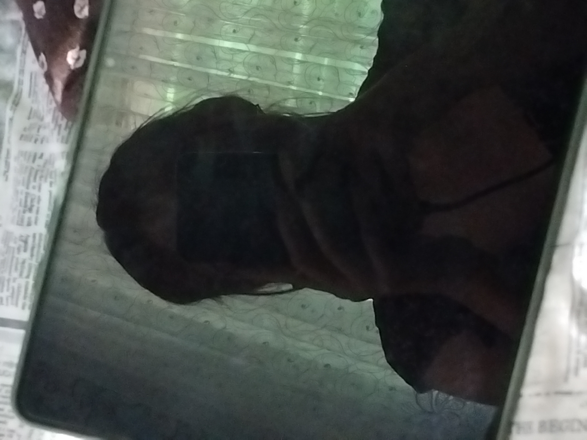 A person is taking a selfie in a dimly lit room. The reflection shows a person holding a smartphone, obscuring their face. The background consists of a curtain with a patterned design and a surface that appears to be covered with newspaper print. The image's lighting is low, creating a silhouette effect. The exact location within Pervozvanivka, Ukraine, cannot be determined from the visual information provided.